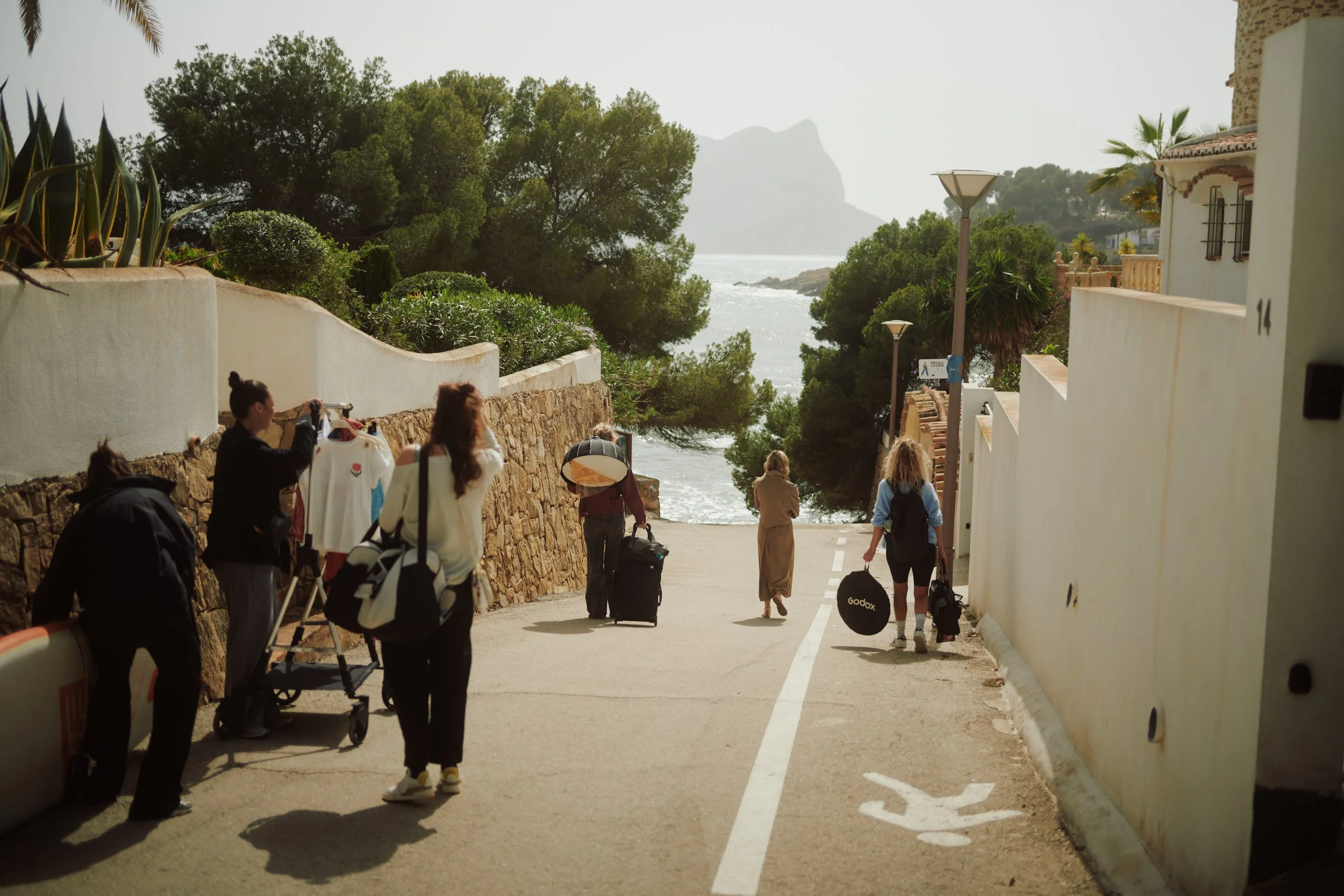 People walking down a street near the coast with luggage and shopping bags, surrounded by greenery and white buildings, with a rocky coastline and mountains in the background.