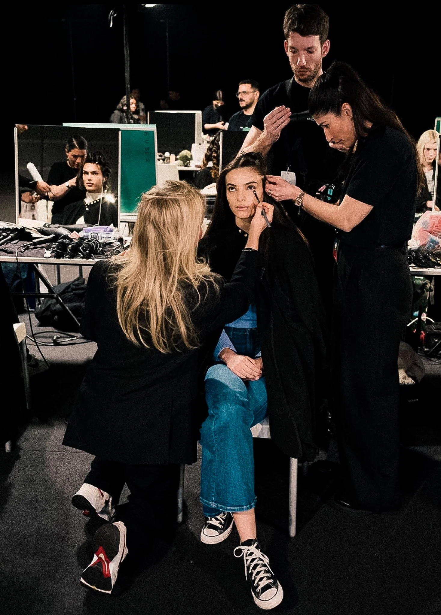 A young woman having her makeup done backstage at a fashion show or photoshoot, with a man standing next to her, and another woman sitting on a chair with her face being painted. The setting is a busy backstage area with tables, mirrors, and makeup a