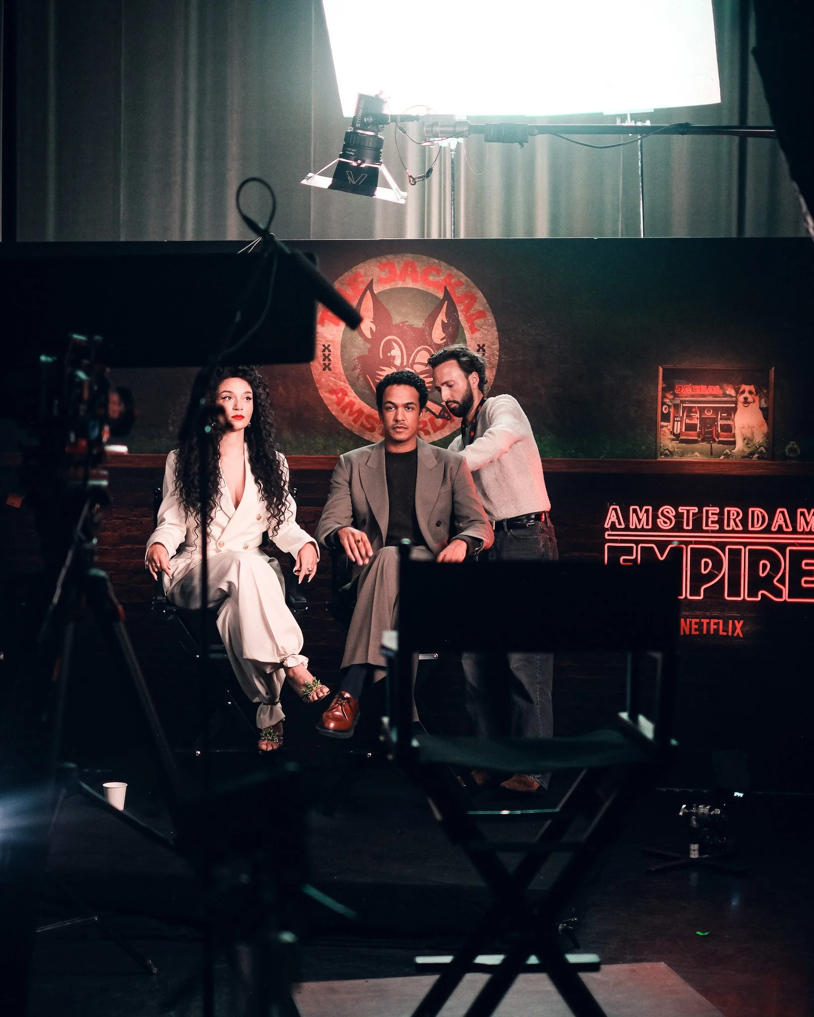 Three people sitting in a studio with a neon sign that says 'Amsterdam Expat'. A woman with curly hair in a white suit, a man in a gray suit, and another man in a light-colored shirt are seated, with one man standing behind the seated man, leaning in