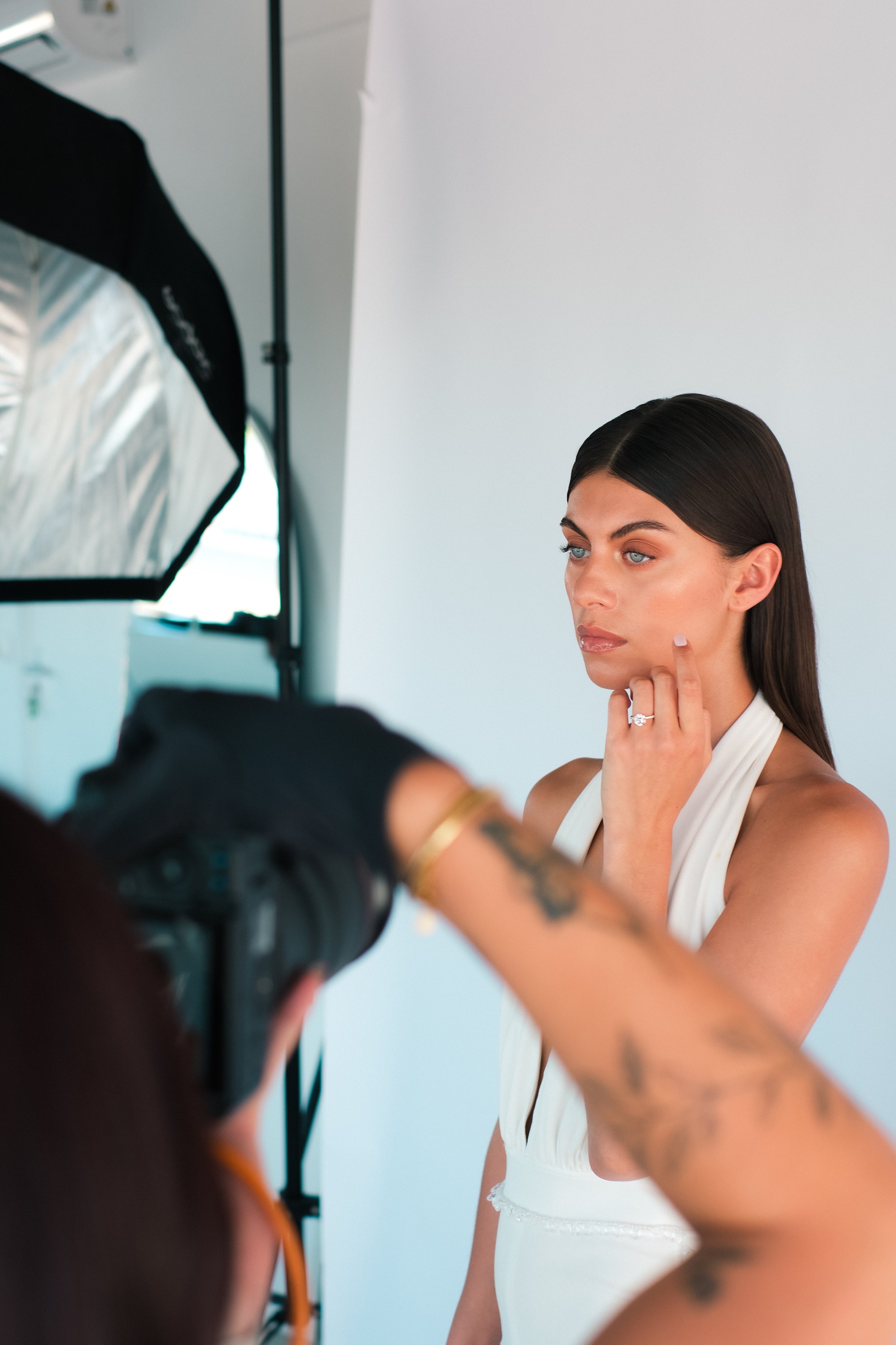 A woman with dark hair, blue eyes, and light makeup posing for a photo in a white dress, while a photographer captures her image with a professional camera and studio lights.