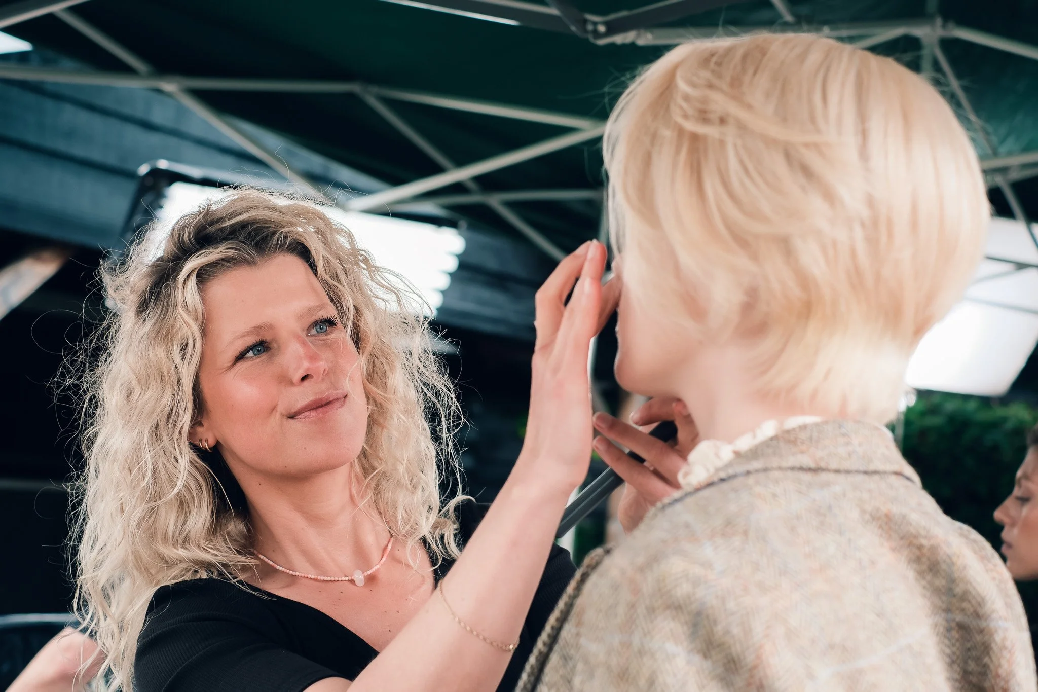 A woman with curly blonde hair and wearing a black top is looking affectionately at another woman with short blonde hair. The woman with short hair is touching her face gently. They are under a canopy with metal framing.