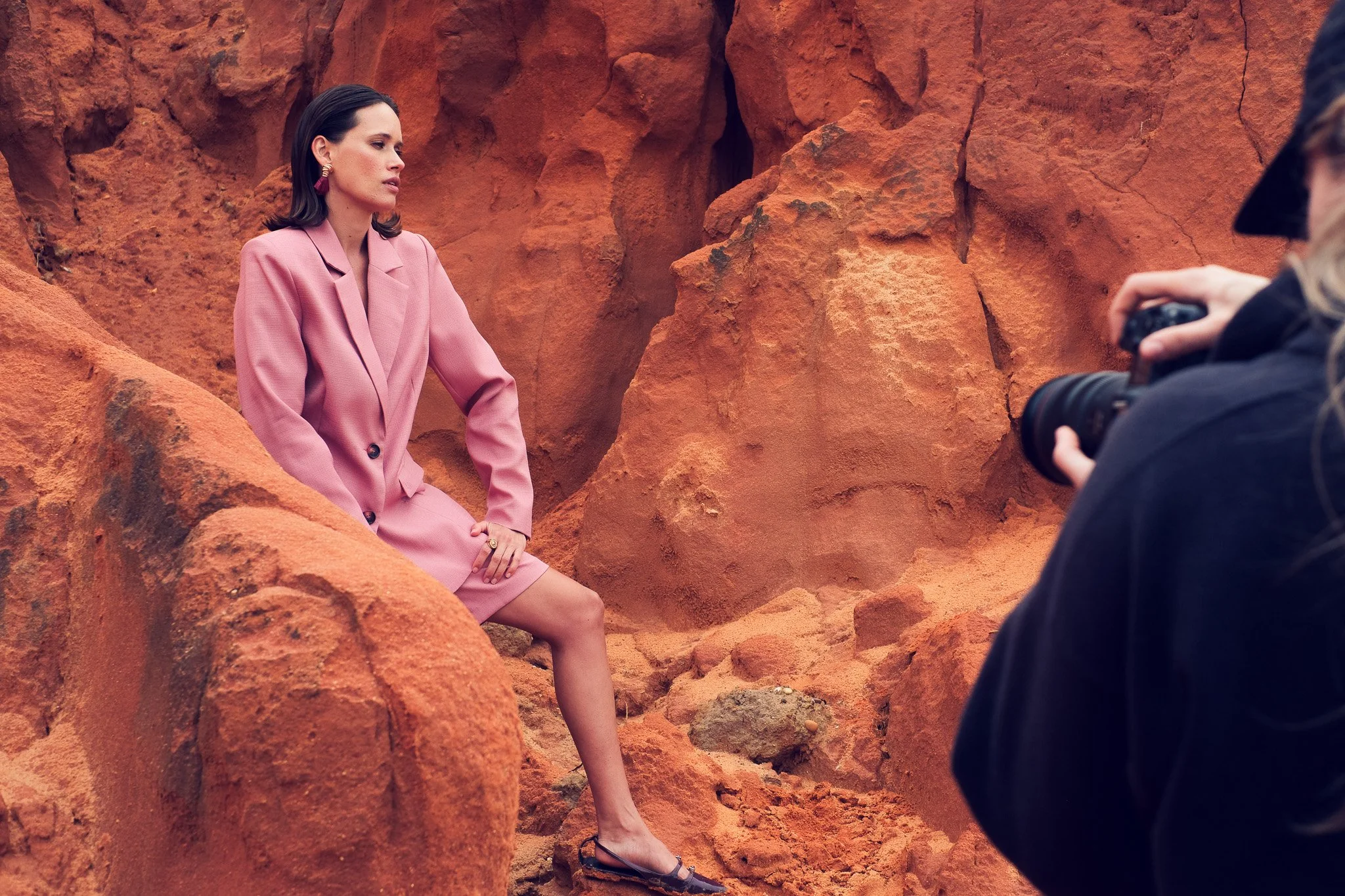 A woman sitting among red rocks during a photo shoot, wearing a pink blazer and skirt, with a photographer capturing her image.