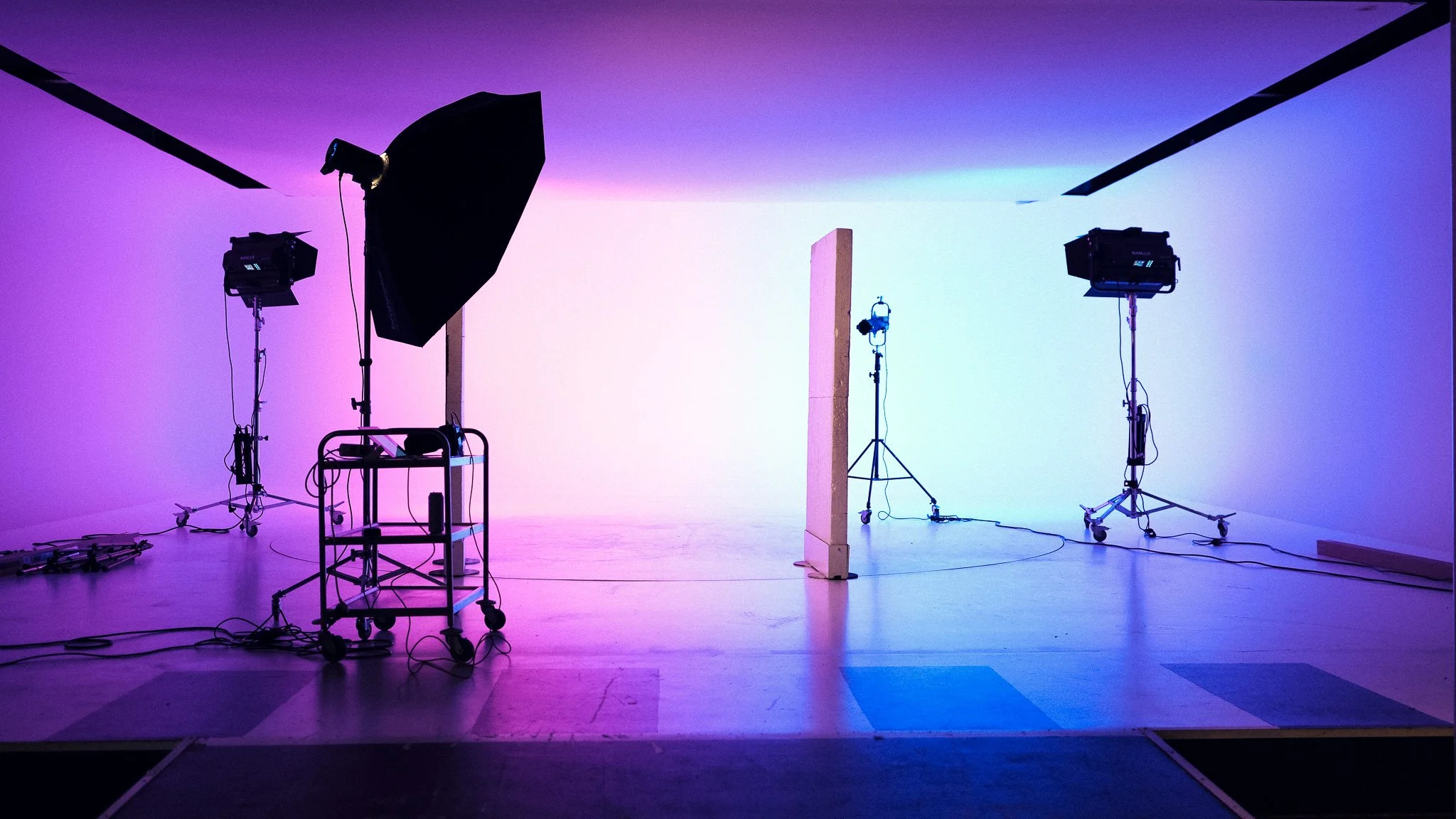 An empty photography studio with professional lighting equipment, a neutral background, and various stands and carts, illuminated with purple and blue lighting.