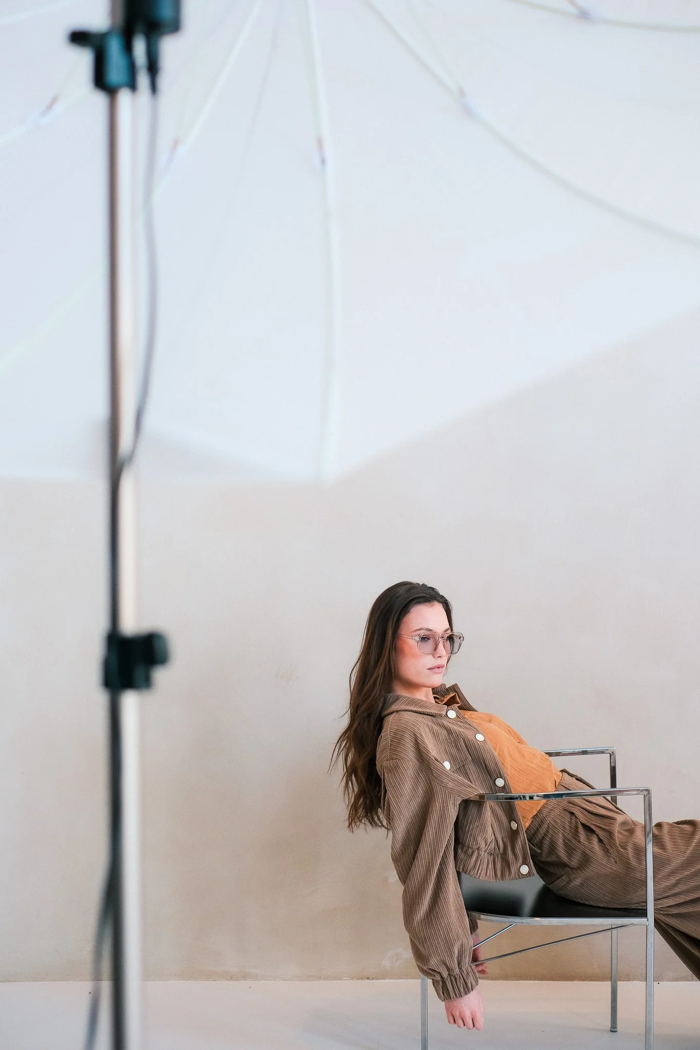A woman with long brown hair wearing glasses and a brown jacket is sitting in a lounge chair against a plain white wall. She has a relaxed expression, and her right arm rests on the chair's armrest while her left arm hangs down.