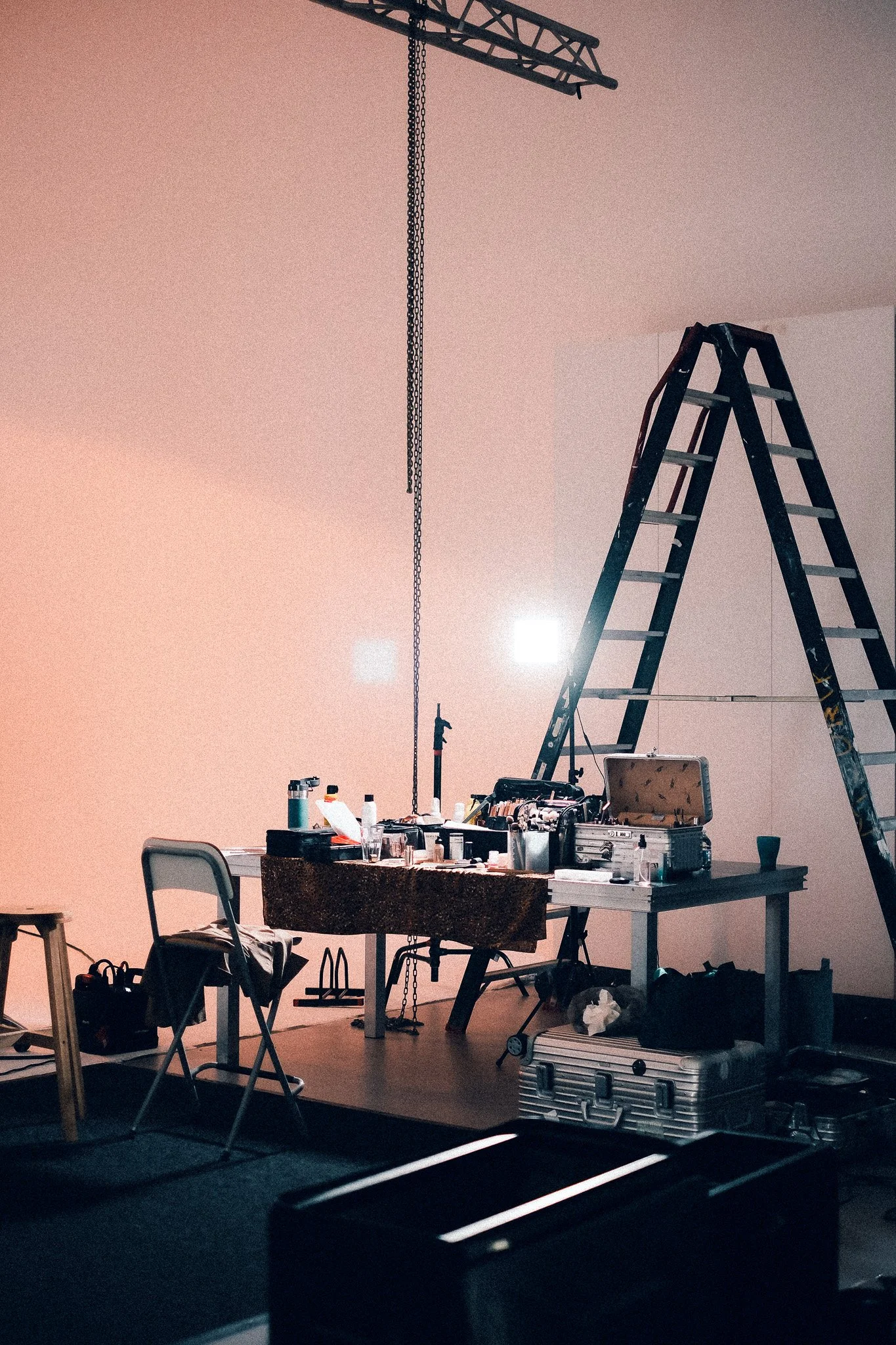 A room with a ladder, table, chairs, and various tools and supplies, suggesting a setup for painting or renovation work. The room has a pinkish wall with a bright light source.
