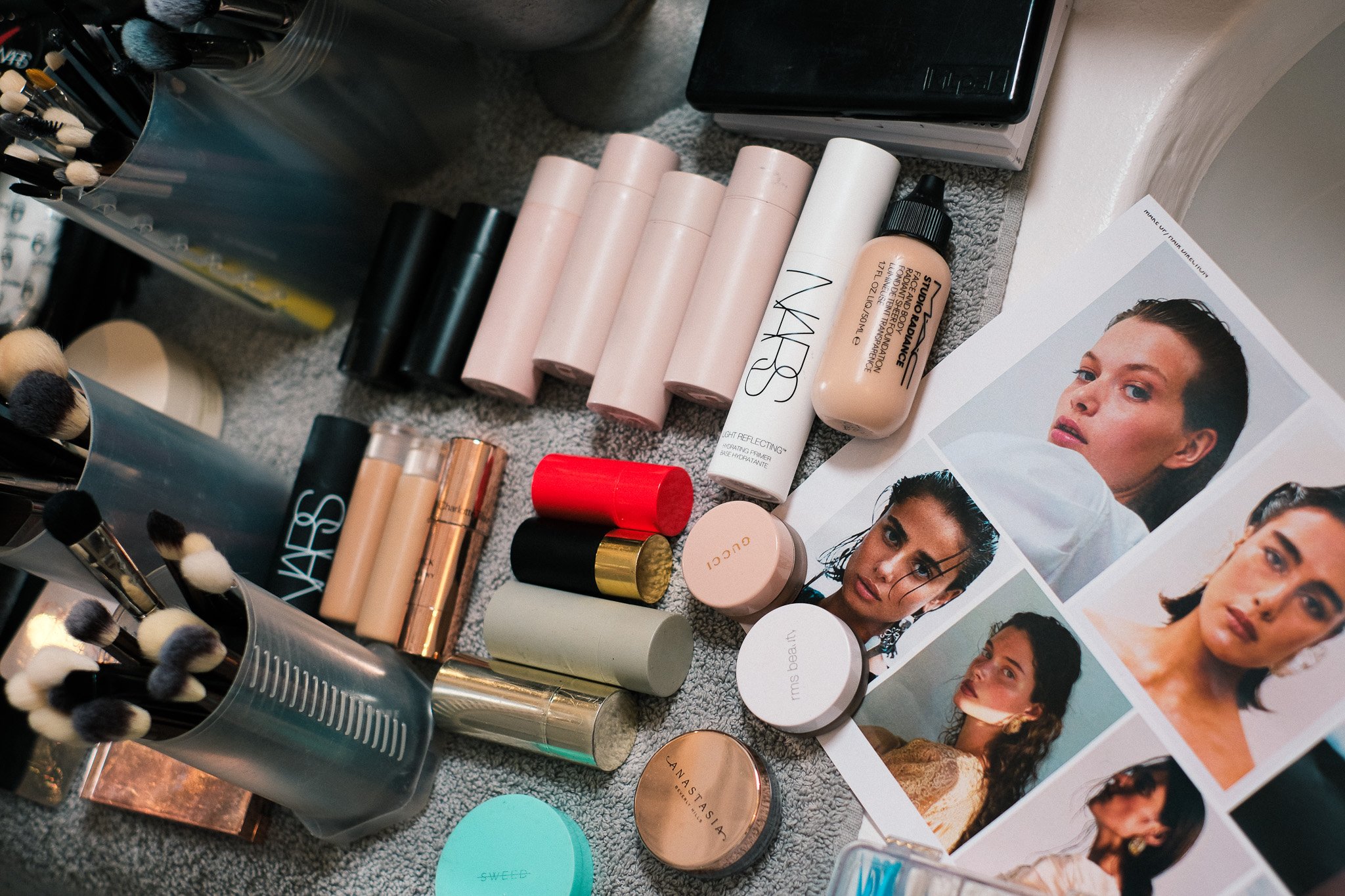 A collection of makeup and skincare products on a grey surface, including foundation bottles, powders in round containers, makeup brushes in a silver holder, and printed images of women with various hairstyles and makeup looks.
