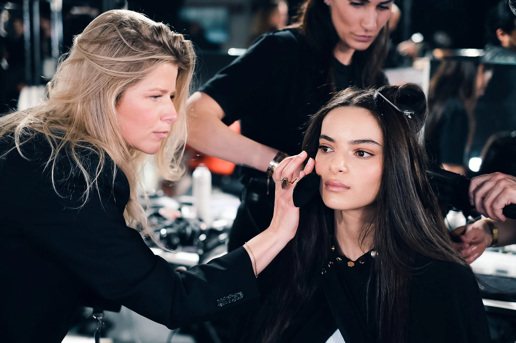 Two women working as makeup artists, one blonde and one brunette, applying makeup to a young woman with dark hair in a busy makeup studio or backstage setting.