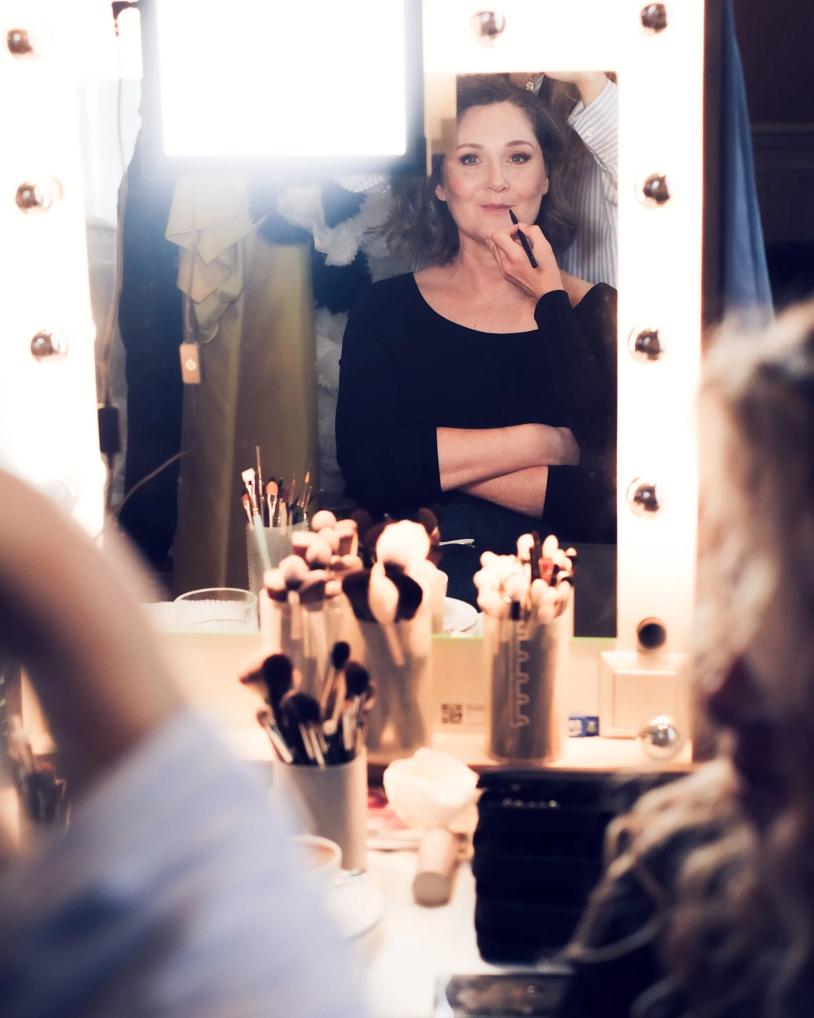 A woman applying makeup in front of a illuminated mirror with various beauty brushes and products on the table.