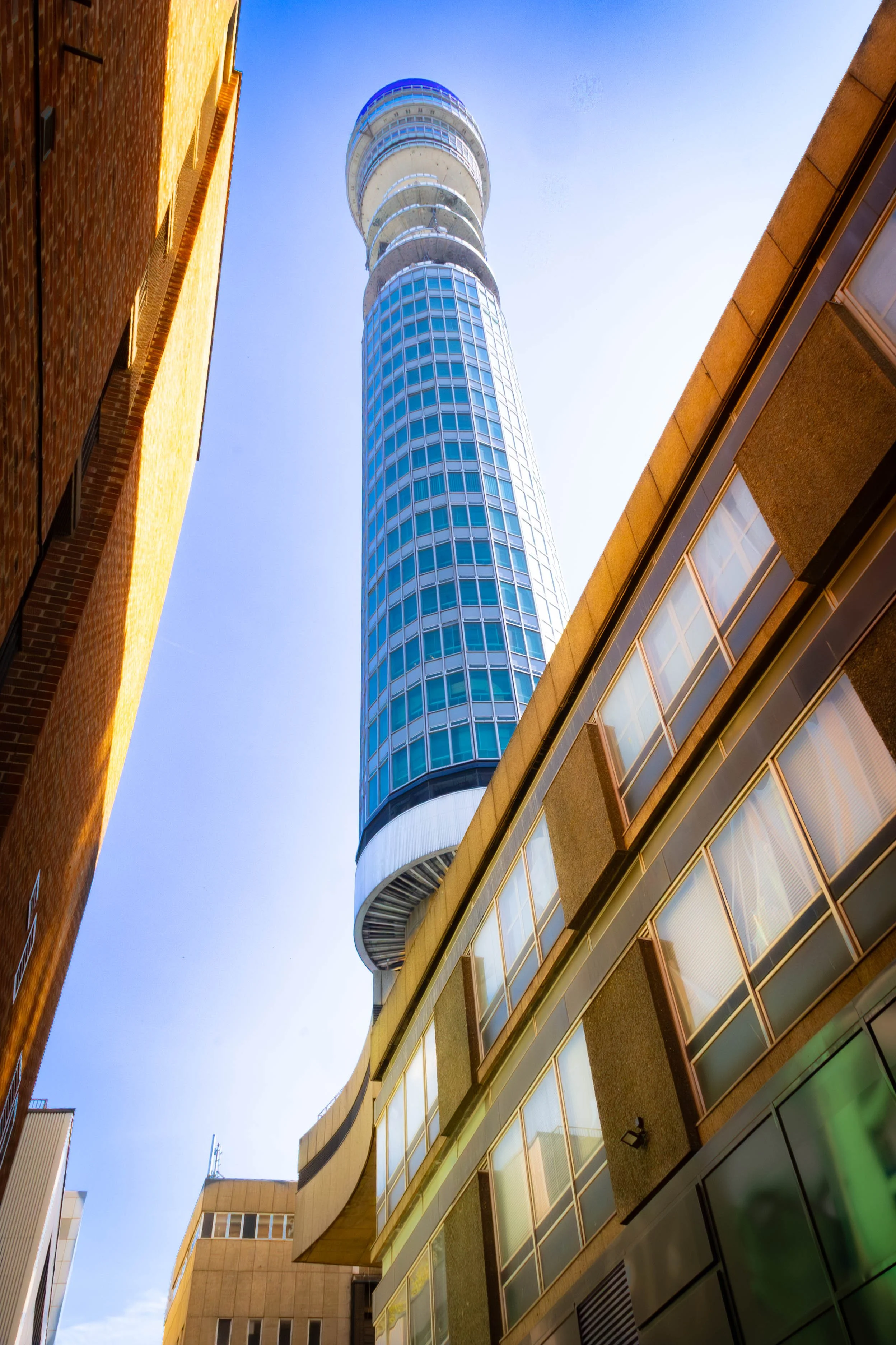 Looking up at a tall, modern glass skyscraper between two older brick buildings on a clear, sunny day.