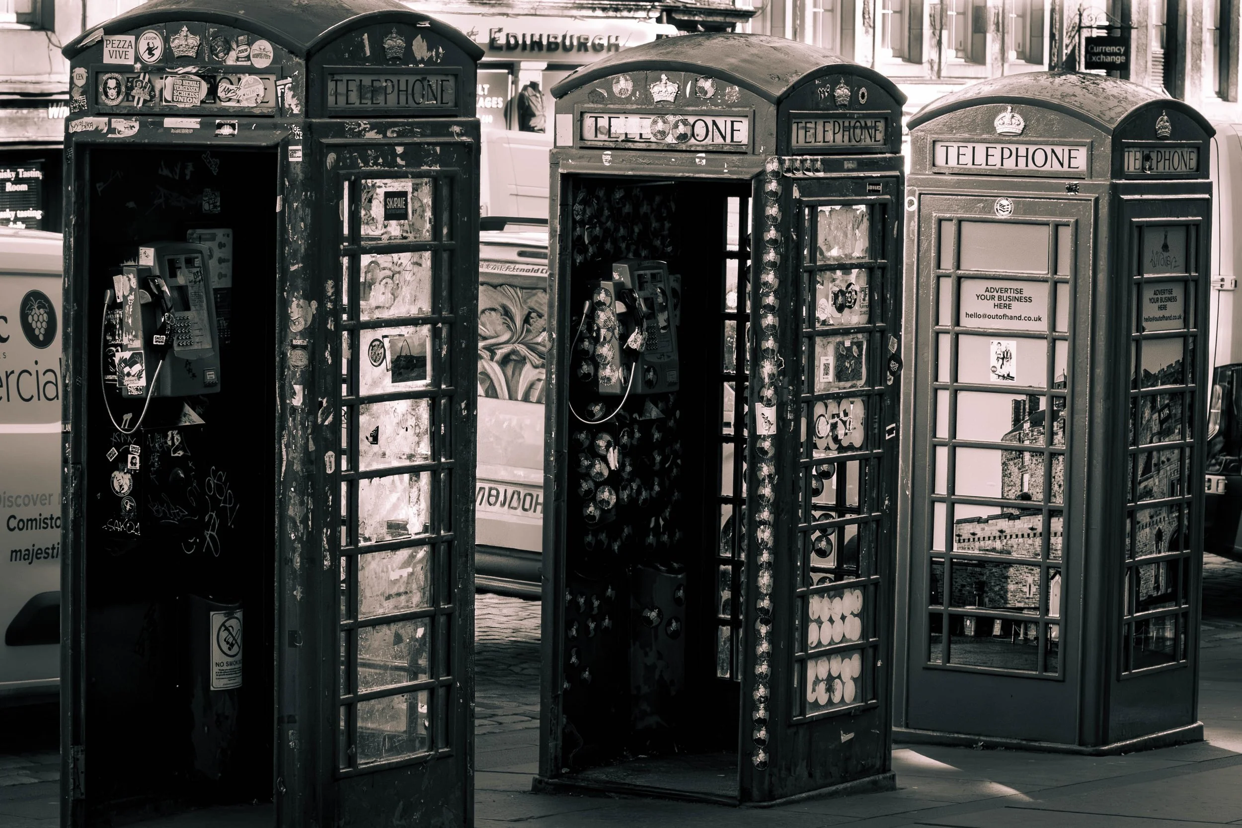 Three vintage telephone booths with weathered exteriors, stickers, and graffiti, situated on a city street with buildings and signs in the background.