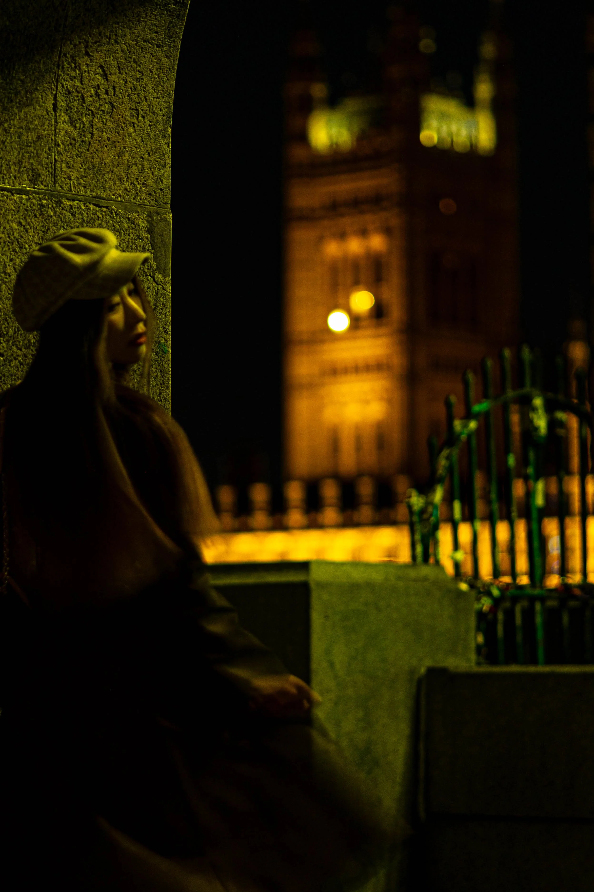 Nighttime scene with a woman wearing a hat, standing against a textured wall on the left, with blurred city lights and a clock tower in the background on the right.