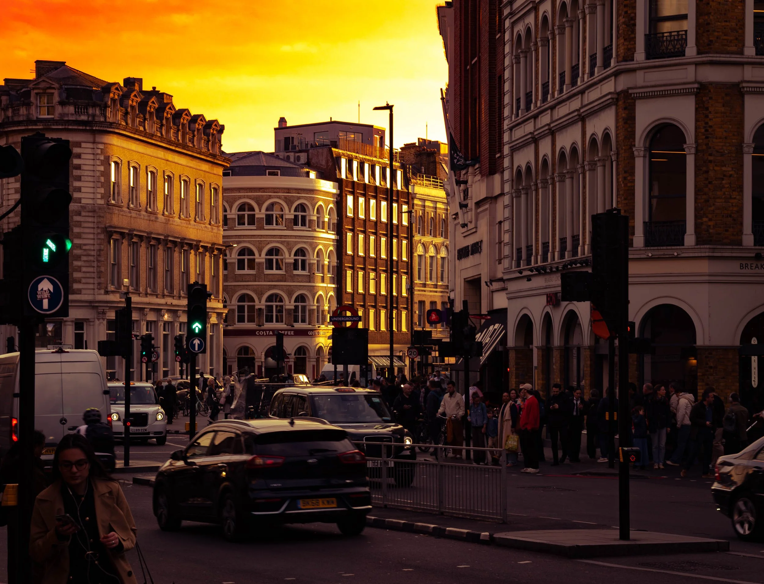 A busy city street at sunset with historic buildings, traffic lights, cars, and pedestrians.