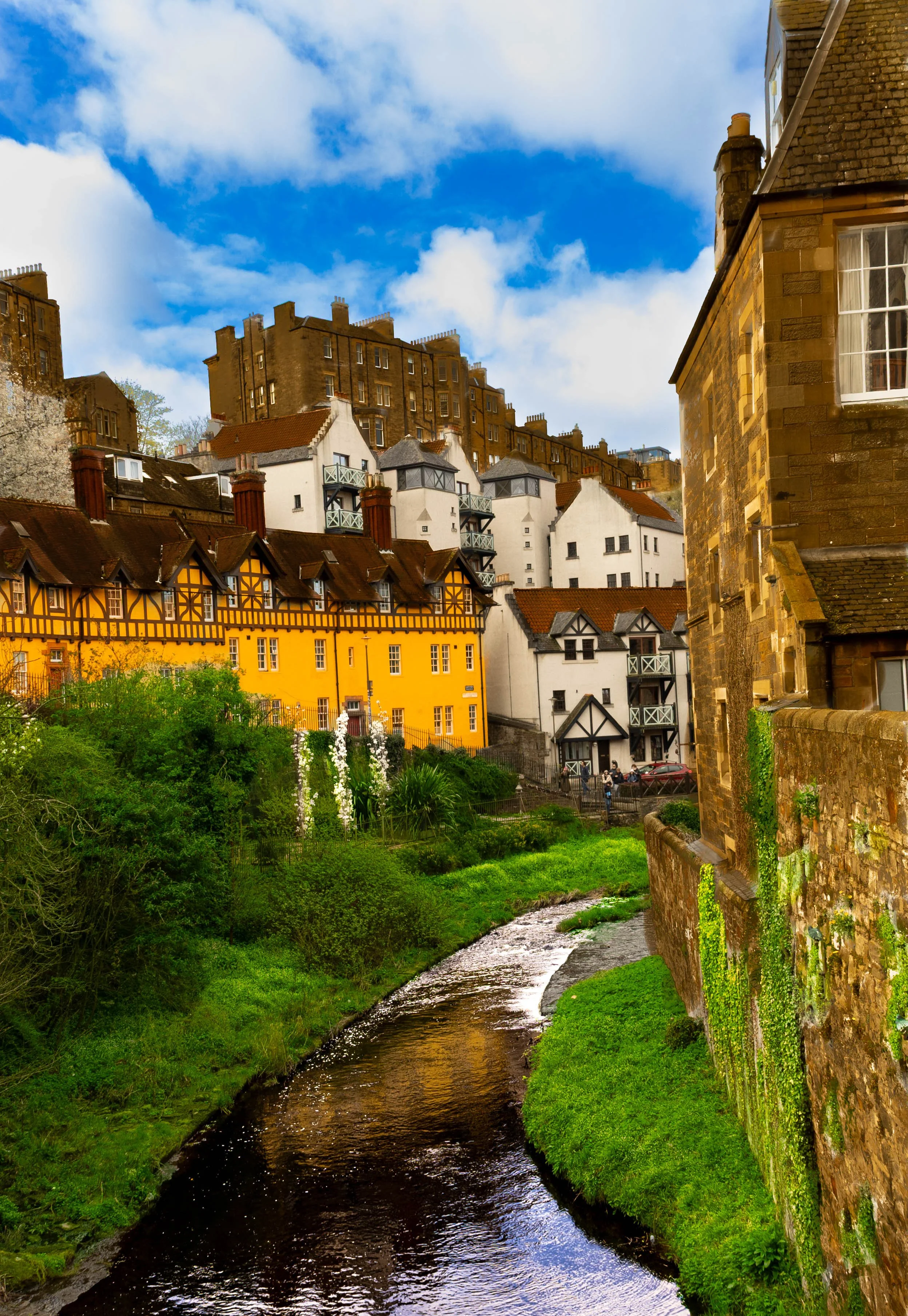 A scenic view of a river flowing alongside colorful houses with a large brown stone building in the background under a partly cloudy sky.