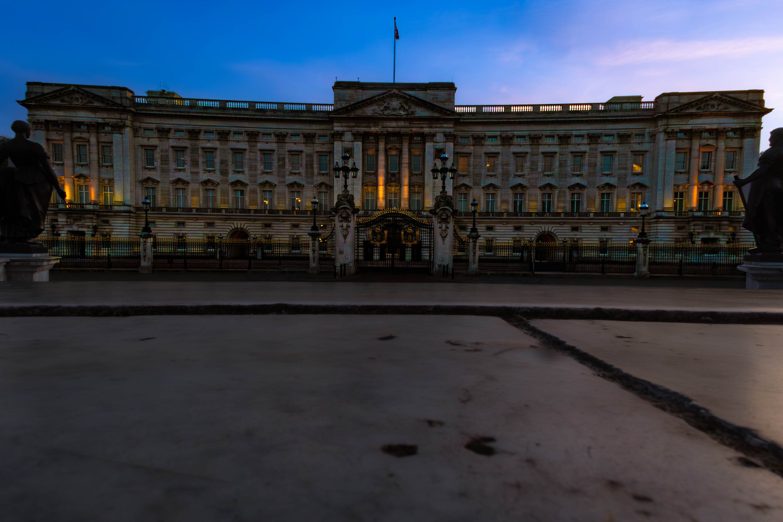 Buckingham palace in twilight with two statues in the foreground, a streetlamp, and a clear sky.