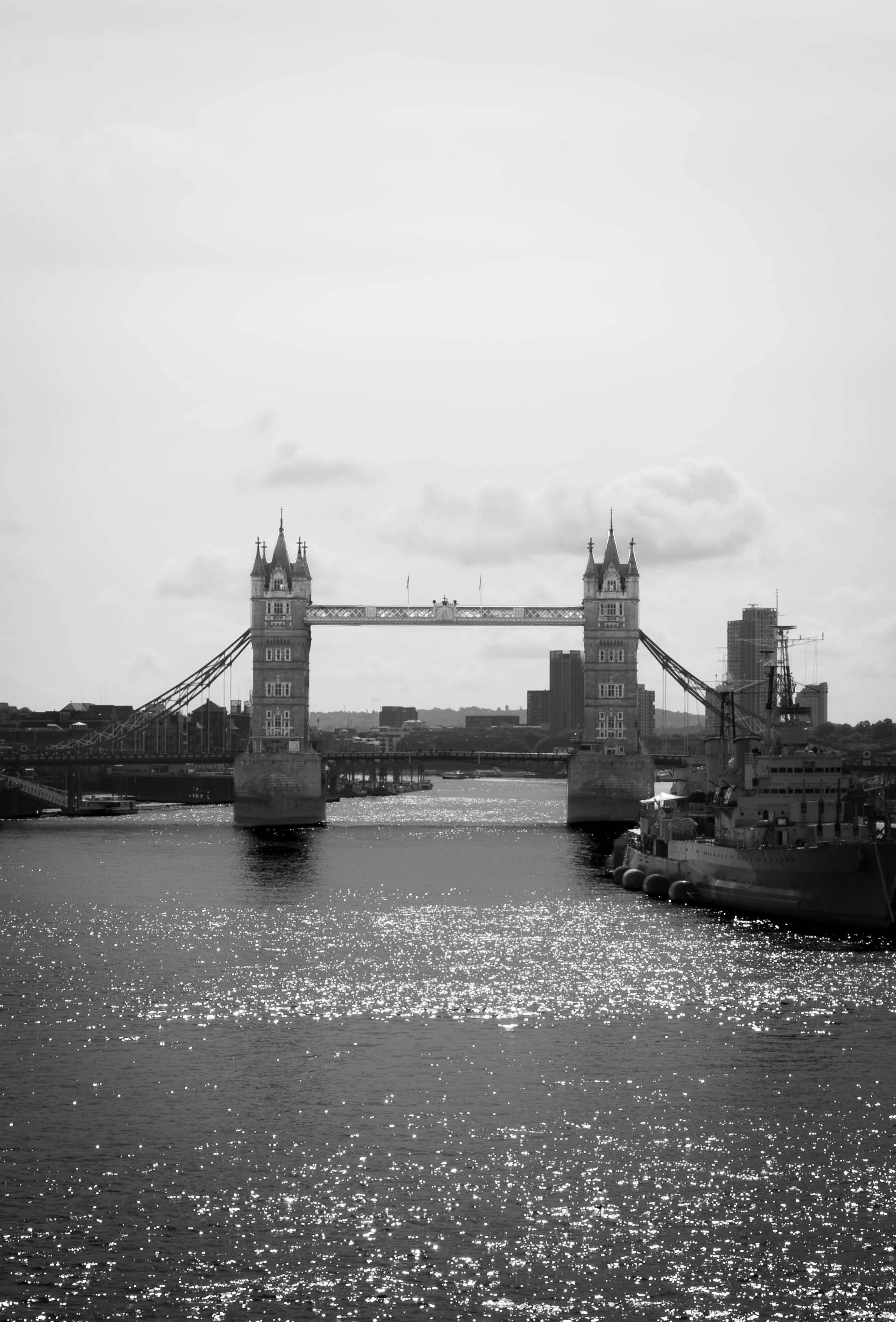 Black and white photo of Tower Bridge in London, England, over the River Thames with boats and buildings in the background.