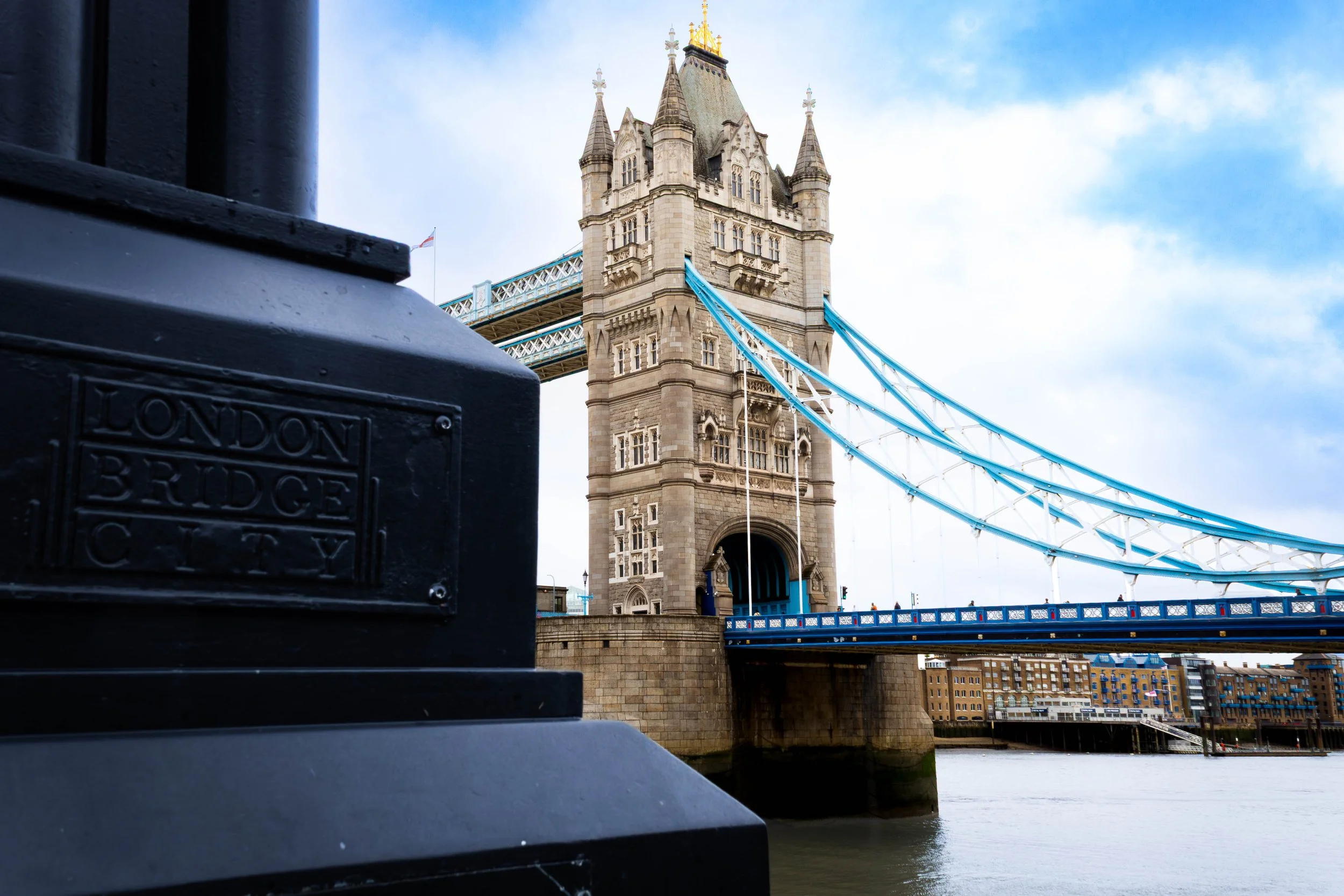 View of London Tower Bridge seen from along the river with some dark foreground elements and a blue sky with some clouds.