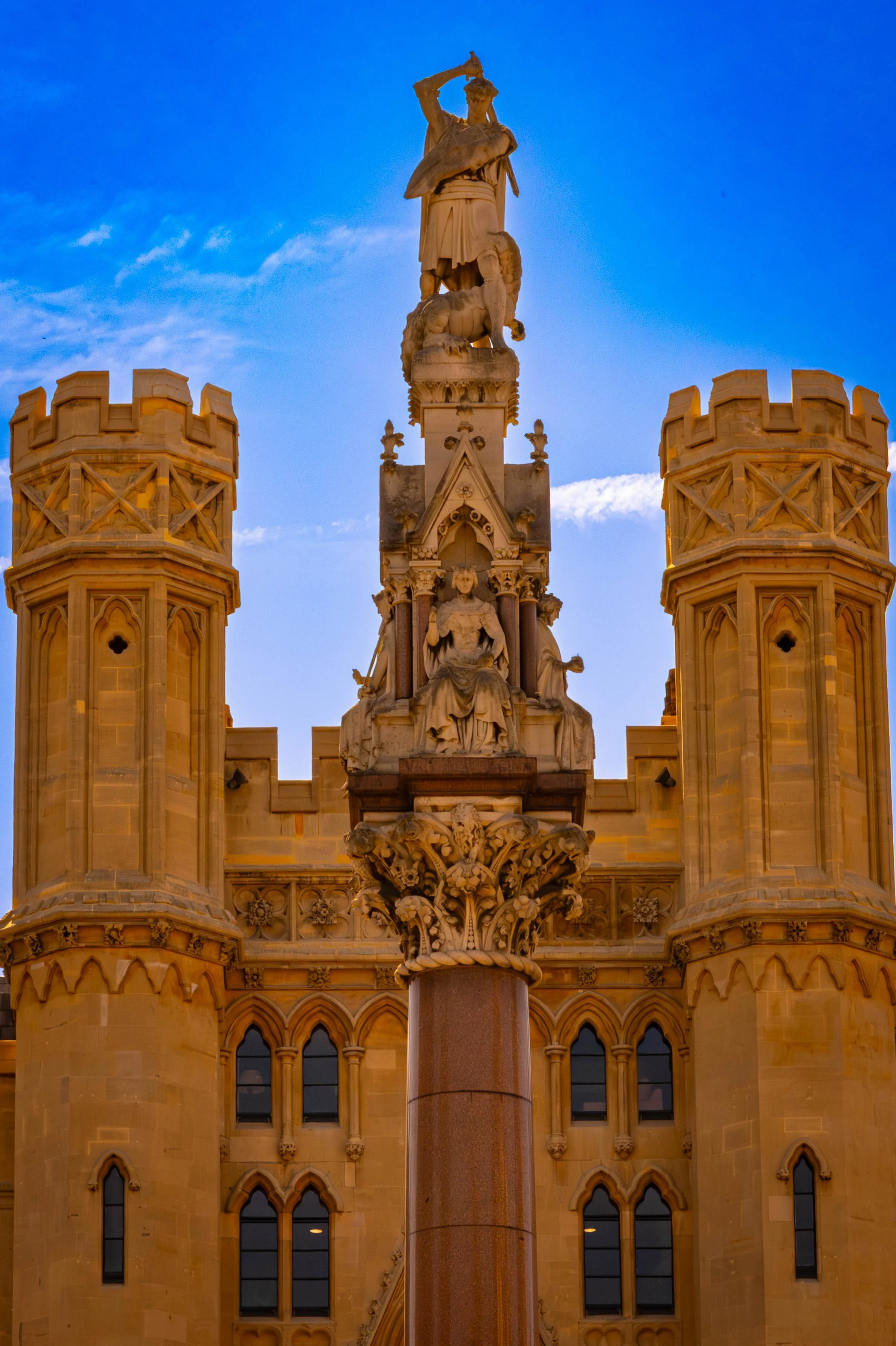A detailed stone monument featuring a sculpted figure of a man holding an object, standing on top of a decorated column with an ornate structure below. Two castle-tower-like structures flank the monument, set against a blue sky with some clouds.