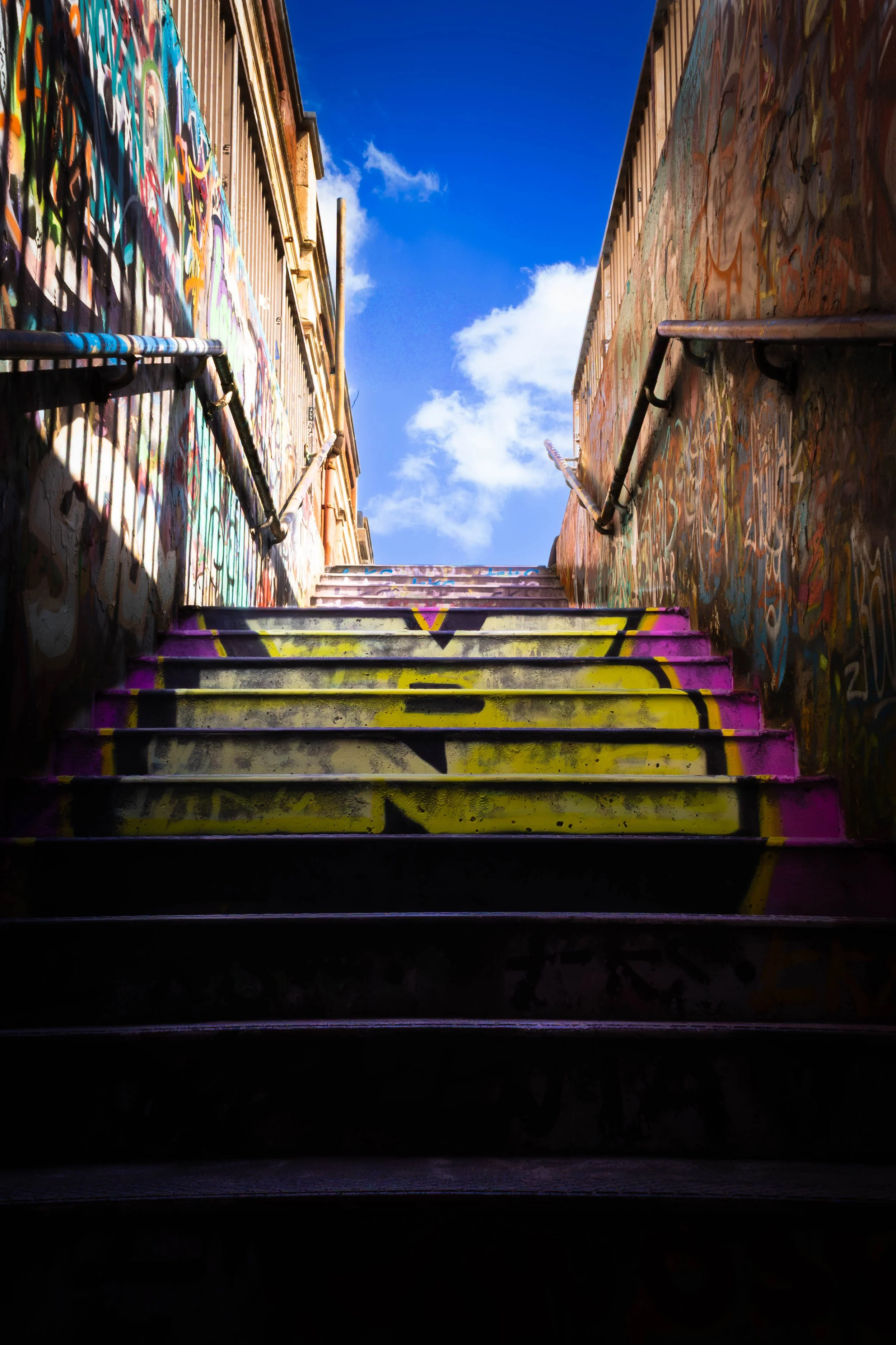 Colorful graffiti on the walls of an alley with a stairway, under a clear blue sky with white clouds.