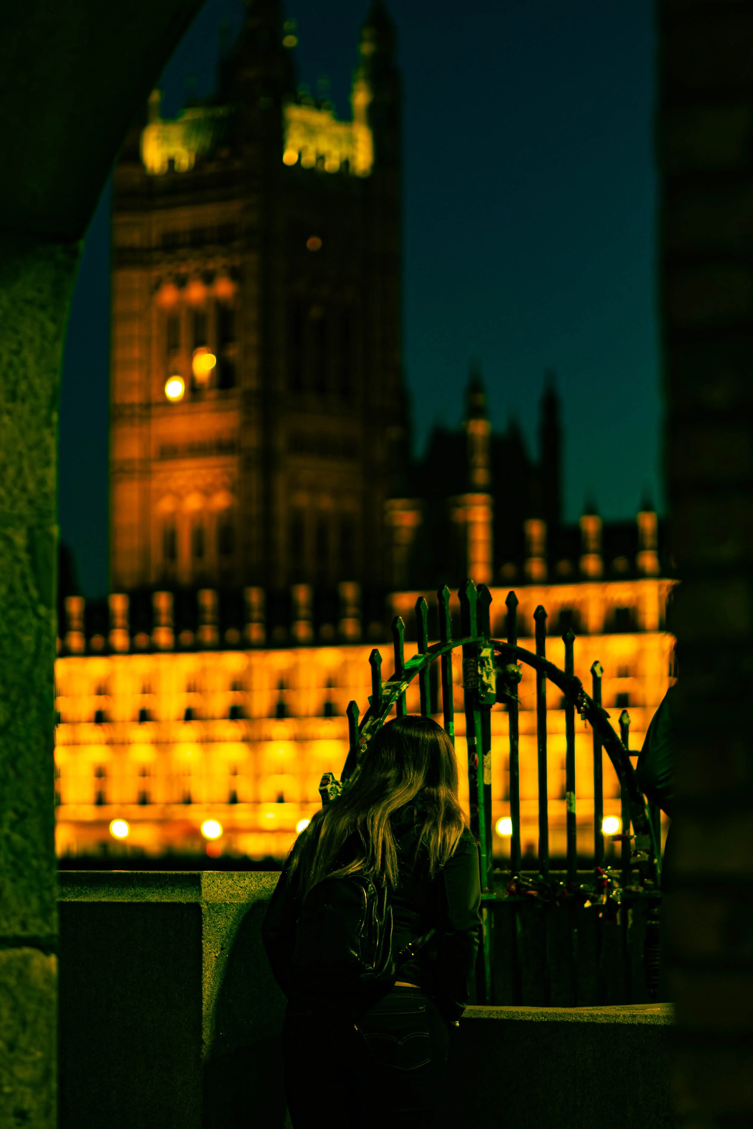 Nighttime view of London's Parliament building, illuminated and blurred in the background, with a women leaning on the stoneworks in the foreground near a metal fence.