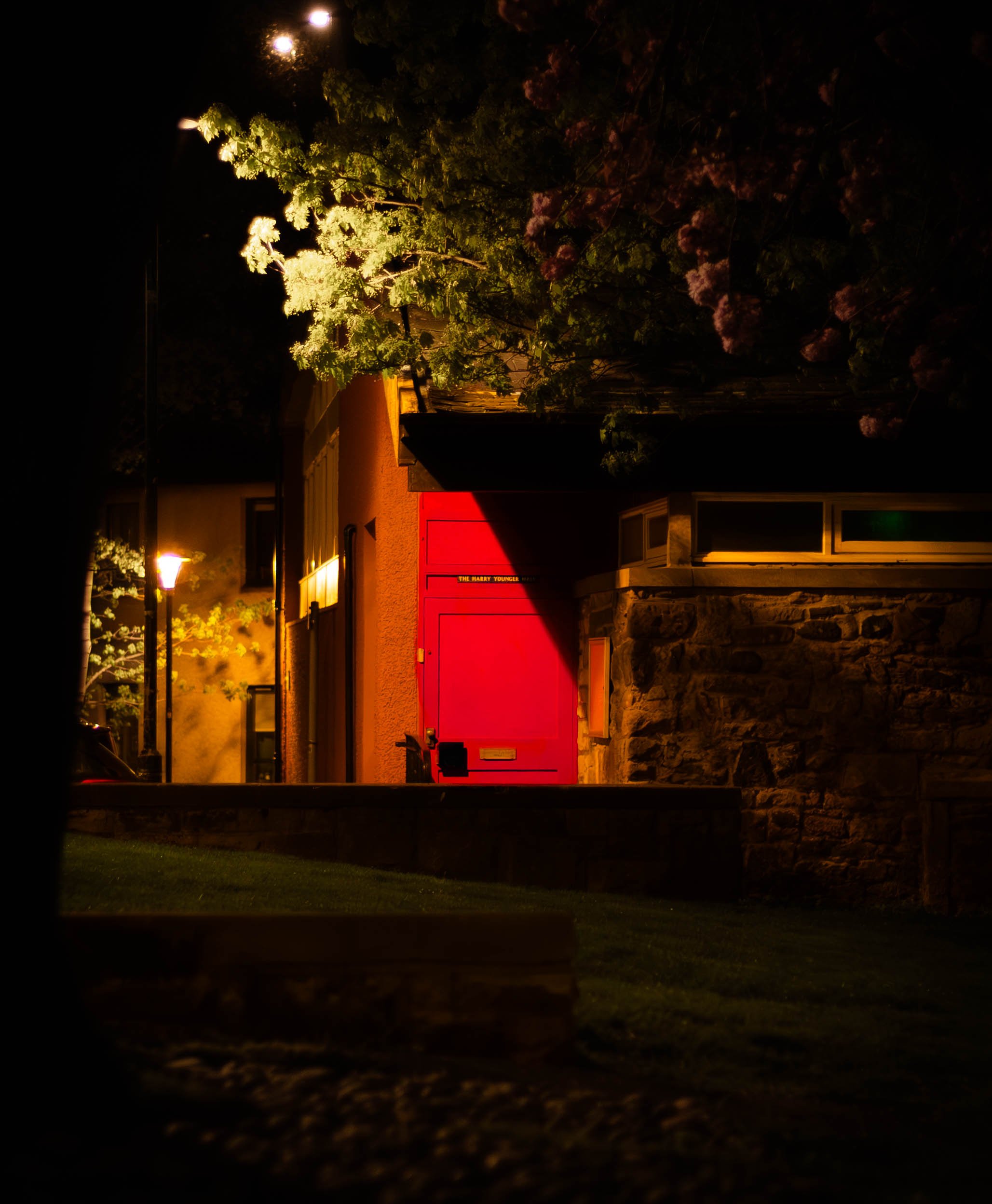 Night view of a small town street with trees illuminated by streetlights, a red building with a sign, and a stone wall in the foreground.