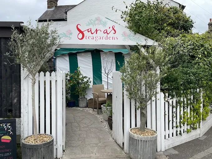 Entrance to Sara's Tea Gardens with white picket fence, potted trees, and outdoor seating under a canopy with green and white stripes.