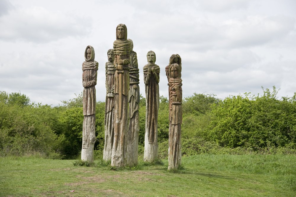 Wooden sculptures of five tall human figures standing outdoors against green trees and cloudy sky.