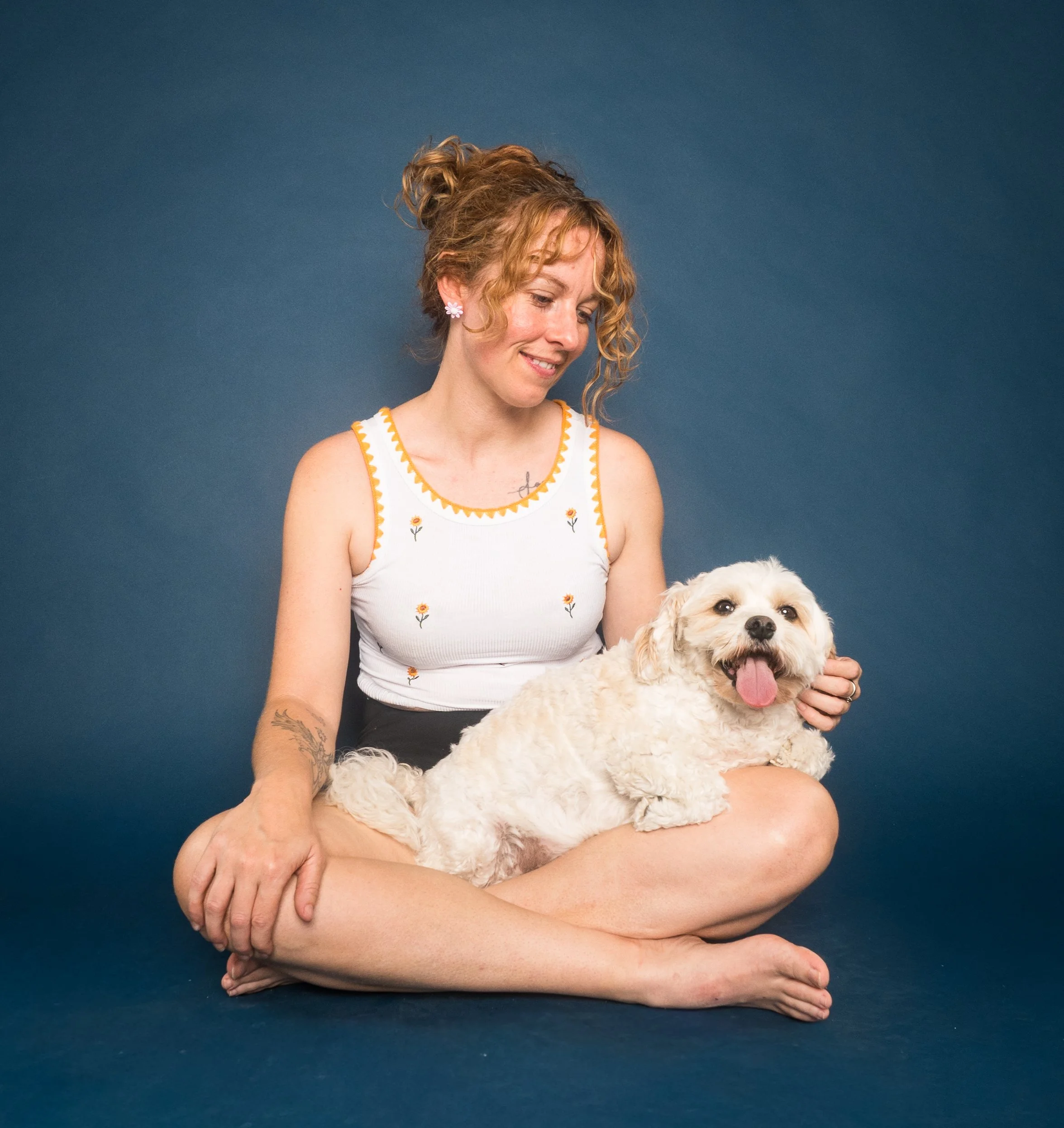 Smiling woman in a floral tank top sitting cross-legged and holding a happy white dog against a dark blue background