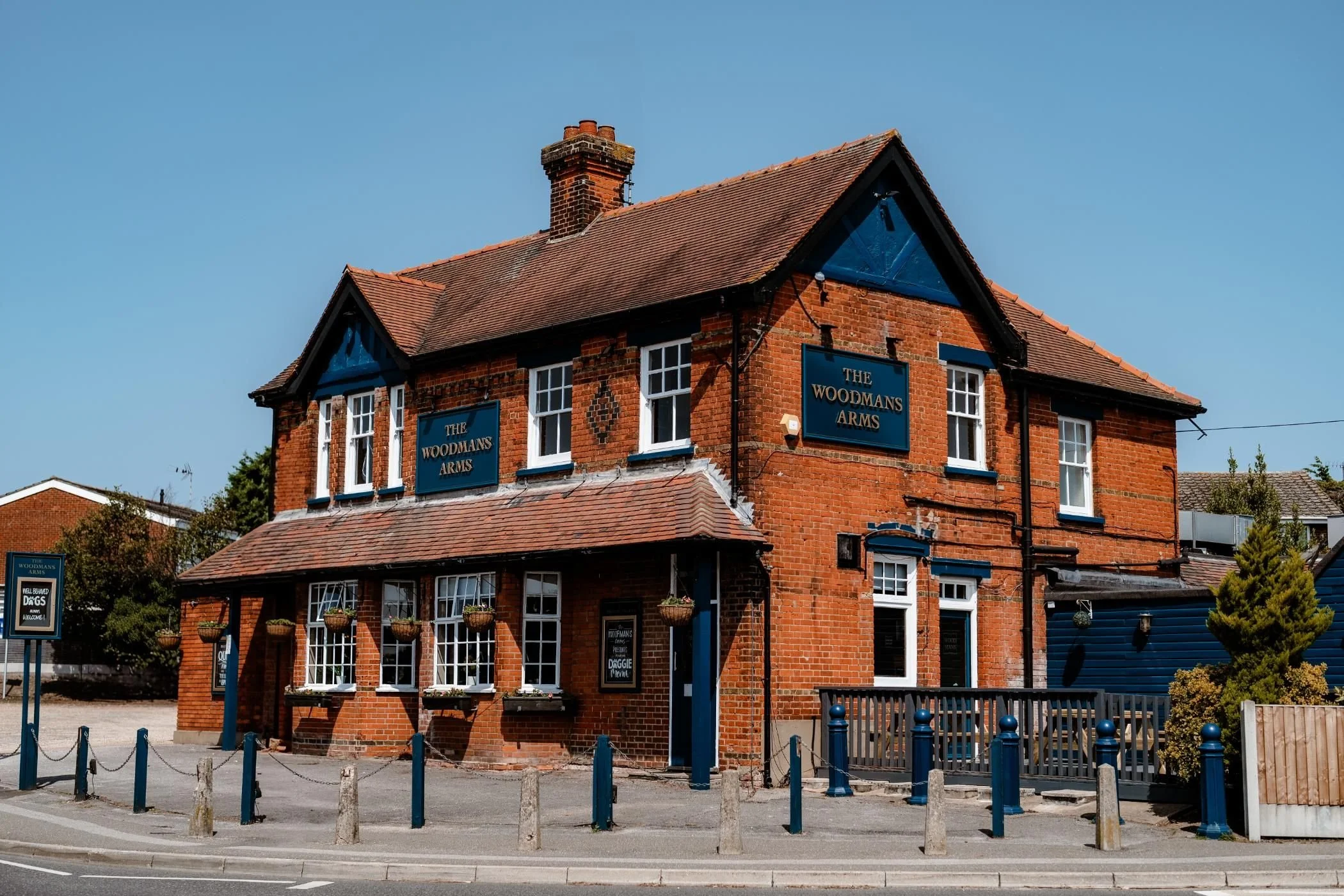 Red brick building with blue accents, multiple windows, and a sign that reads 'The Woodmans Arms.' There are several hanging flower baskets on the front, and a fenced outdoor area with blue posts in the front. Clear blue sky in the background.