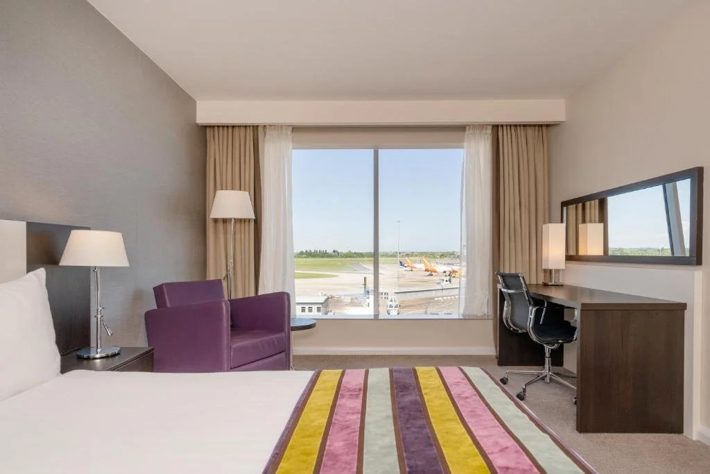 A hotel room with a bed, purple armchair, desk with a chair, large window with curtains showing an airport runway and planes outside.