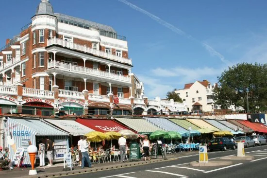 Street view with a large red brick hotel and storefronts with colorful awnings, outdoor seating, and pedestrians.