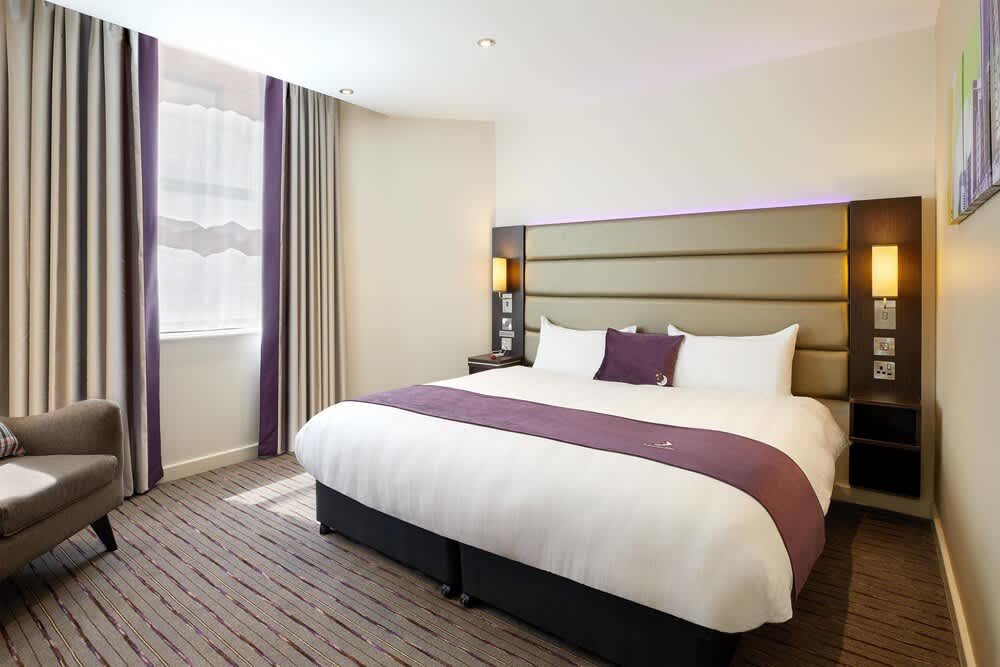Hotel room with a large bed, beige and purple headboard, purple decorative pillow, white linens, bedside lamps, window with beige and purple curtains, and a small beige sofa.