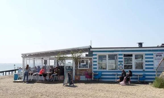 A beachside cafe with blue and white striped exterior, outdoor seating, and people sitting and standing by the water.