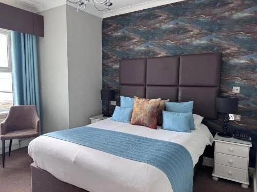 A hotel room with a large bed, blue bed runner, and multiple pillows, a padded headboard, side tables with lamps, a chair near the window, and a wall with a colorful abstract mural.