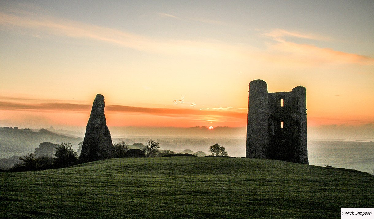 Sunset over rolling hills with ruins of an ancient castle and a tower in the foreground.