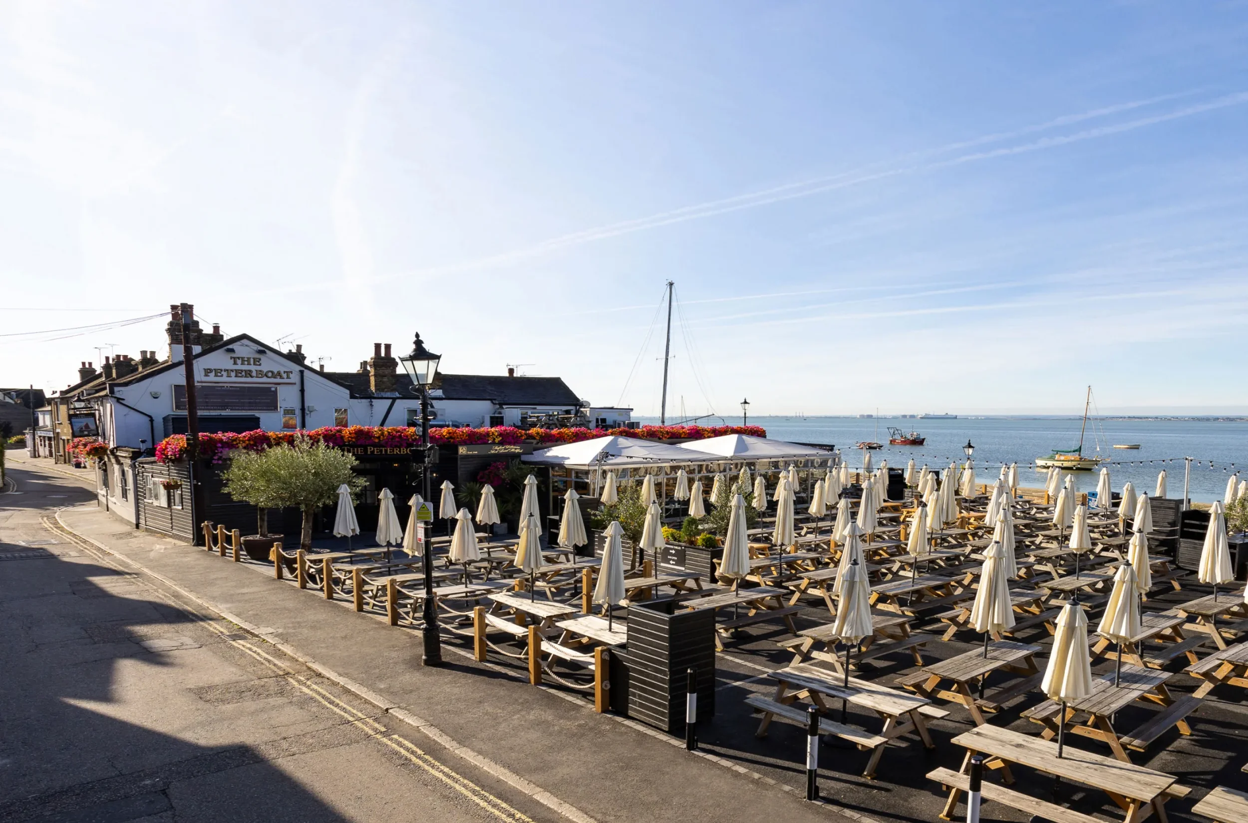 A seaside restaurant with outdoor seating, umbrellas, and a view of the water and boats, with a sign reading 'The Peterboat' on the building.