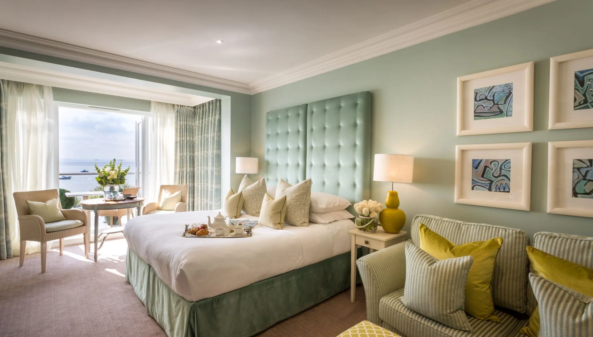 Bright hotel room with large window open to water view, king-sized bed with green upholstered headboard, bedside lamps, armchairs with yellow pillows, and artwork on the wall.