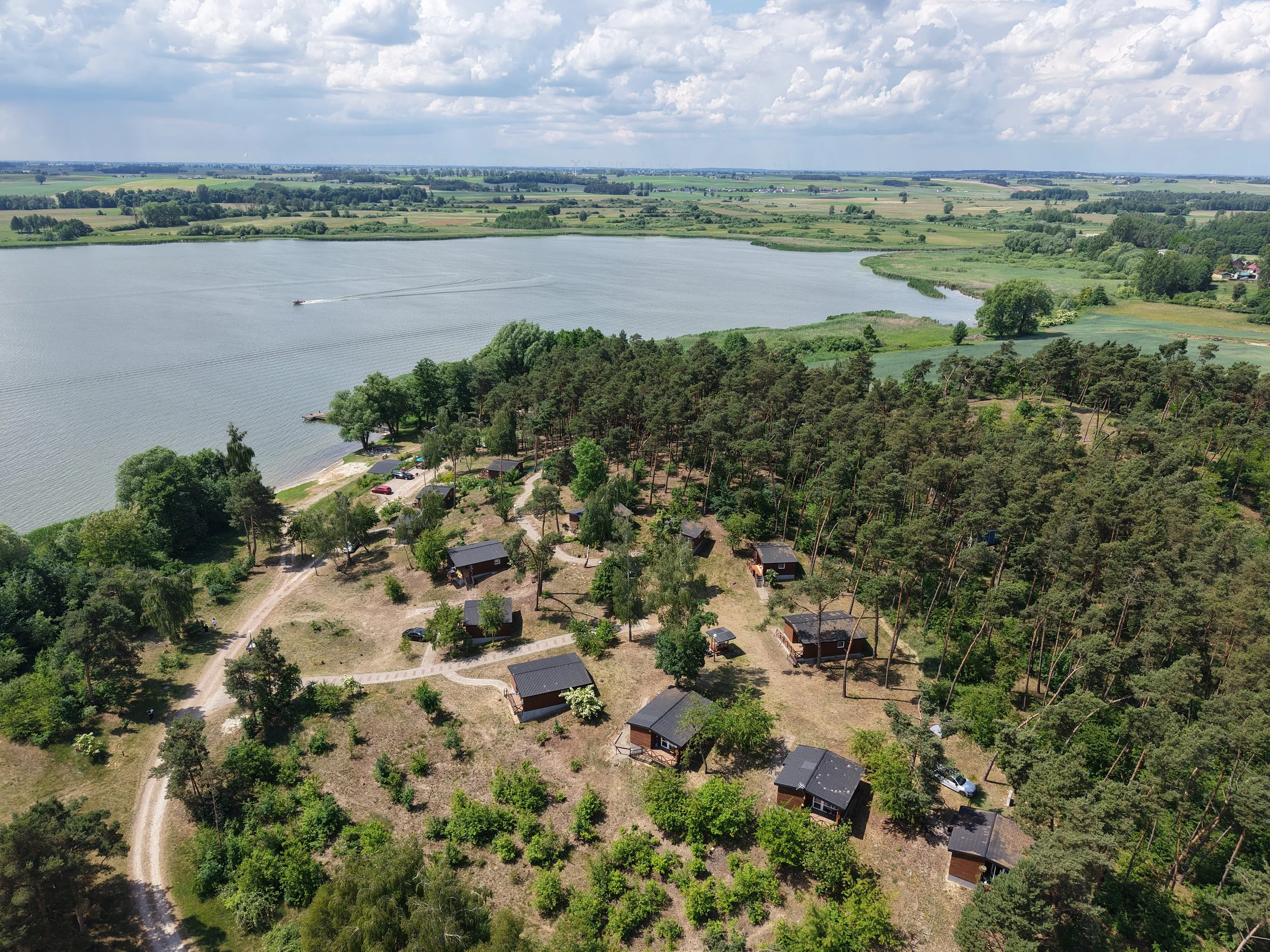 Widok z lotu ptaka na jezioro Wolickie w Pturku z łódką na wodzie, otoczone polami i drzewami, dokami letniskowymi.