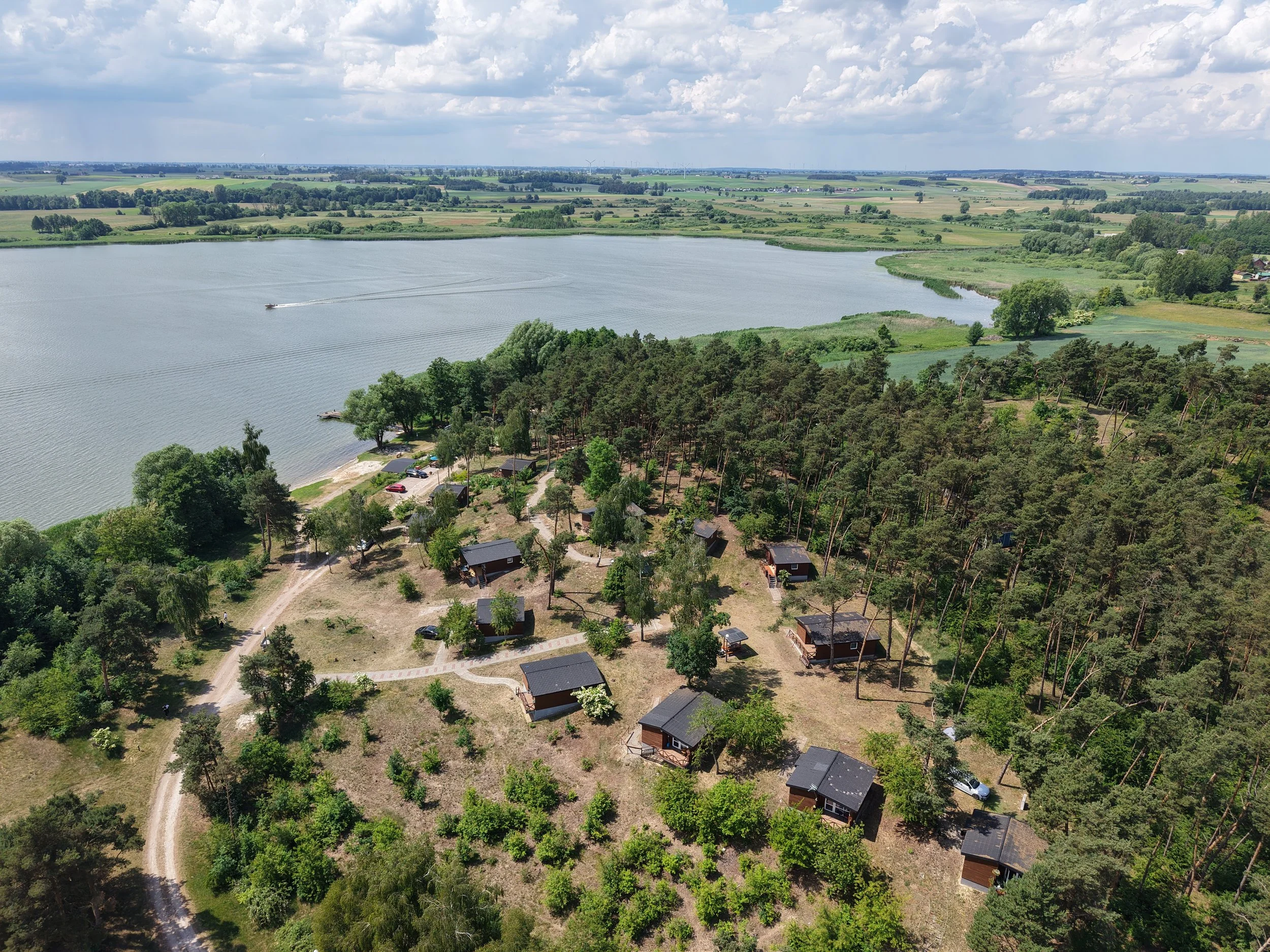 Widok z lotu ptaka na ośrodek wypoczynkowy nad jeziorem w Wolickim w kujawsko-pomorskim województwie. Domki położone nad jeziorem. Drewniane domki wśród drzew, piaszczysta plaża i łódź na wodzie,  woda, zielone pola, las, wakacje, weekend.