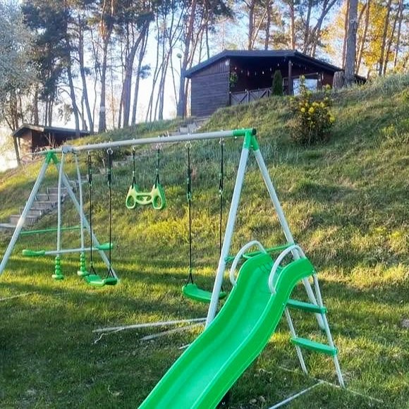 Zielony plac zabaw ze zjeżdżalnią, huśtawkami i zestawem huśtawek dla dzieci na trawiastym podwórku z drzewami i domem w tle nad jeziorem w Pturku, w ośrodku wypoczynkowym nad jeziorem Wolickim.