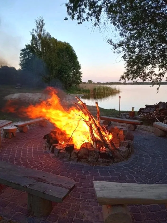 Ognisko płonące w pobliżu brzegu jeziora Wolickiego w Pturku, otoczone drewnianymi ławkami i cegłami, z drzewami i wodą w tle. Palenisko nad jeziorem na plaży. Relaks i wypoczynek.