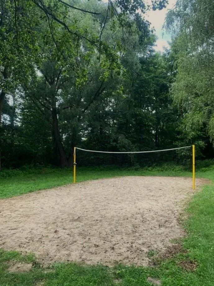 Boisko do siatkówki plażowej z siatką, otoczone zieloną trawą i drzewami nad jeziorem Wolickim w Pturku w ośrodku wypoczynkowym