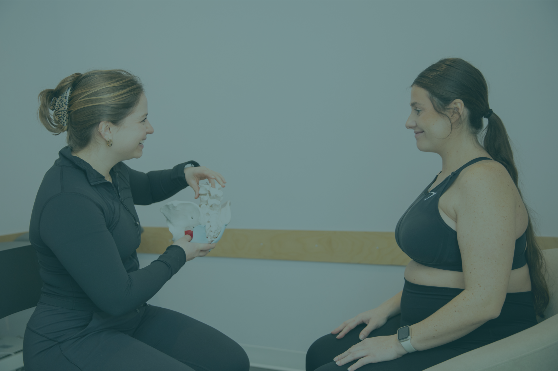 Une femme en uniforme de femme médecin ou kinésithérapeute présente un modèle d'anatomie pelvienne à une femme en vêtements de sport, dans une salle d'examen ou de consultation.
