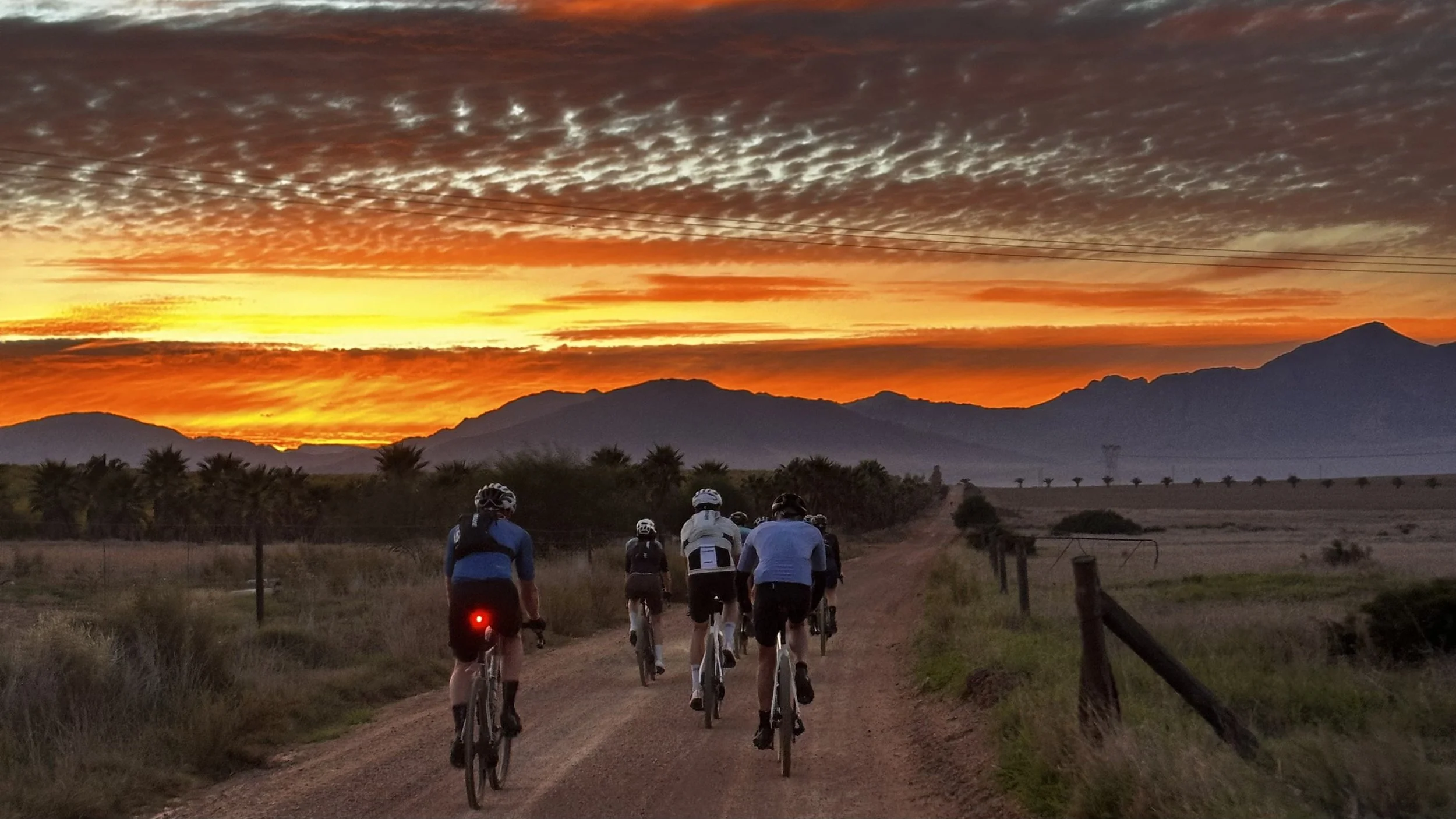 A group of cyclists riding on a dirt trail during sunrise with mountains and a colorful sky in the background.