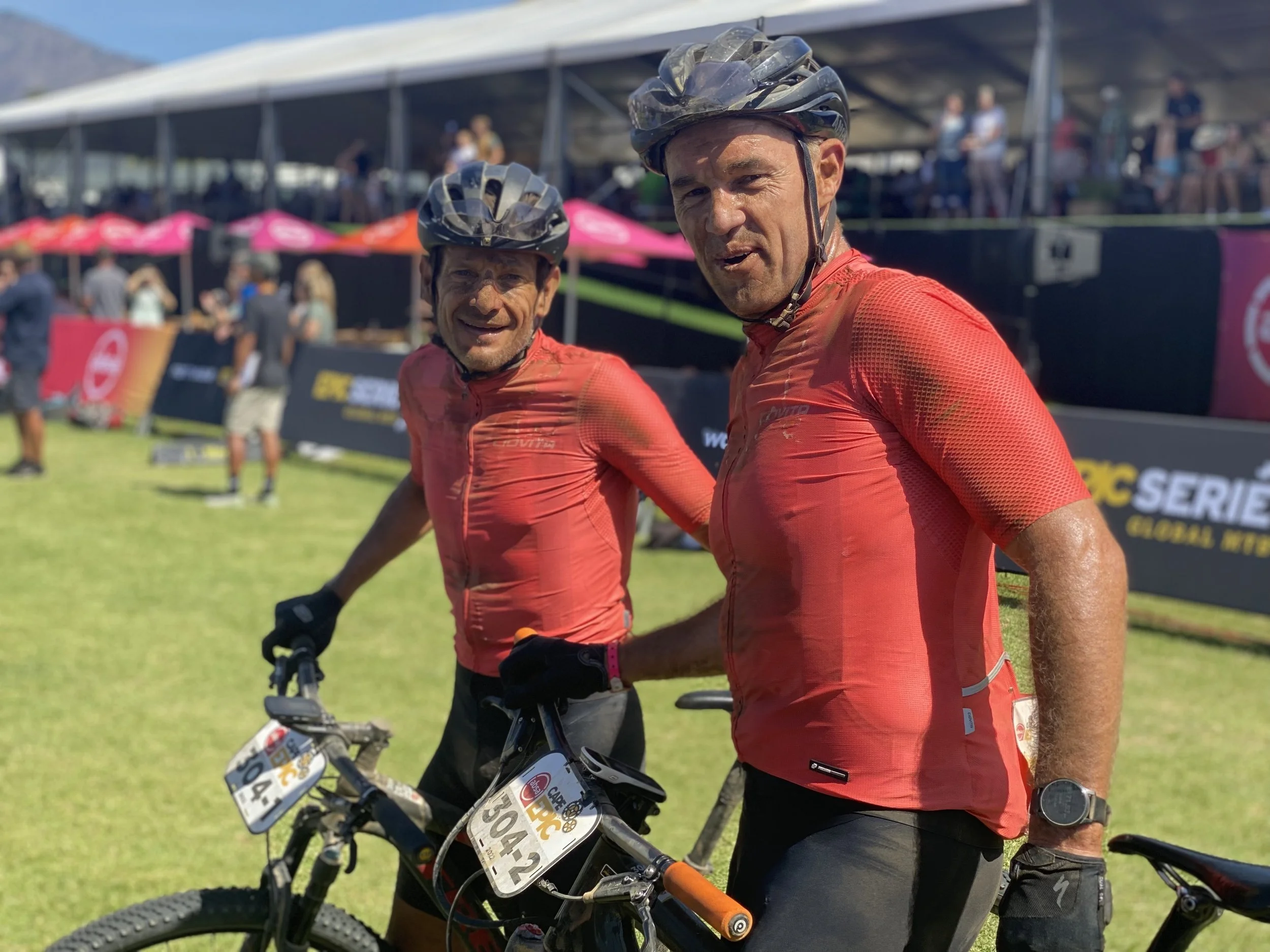 Two male mountain bikers wearing red cycling jerseys and black helmets at the Cape Epic finish, with bikes in front of them, and a crowd and event tents in the background.