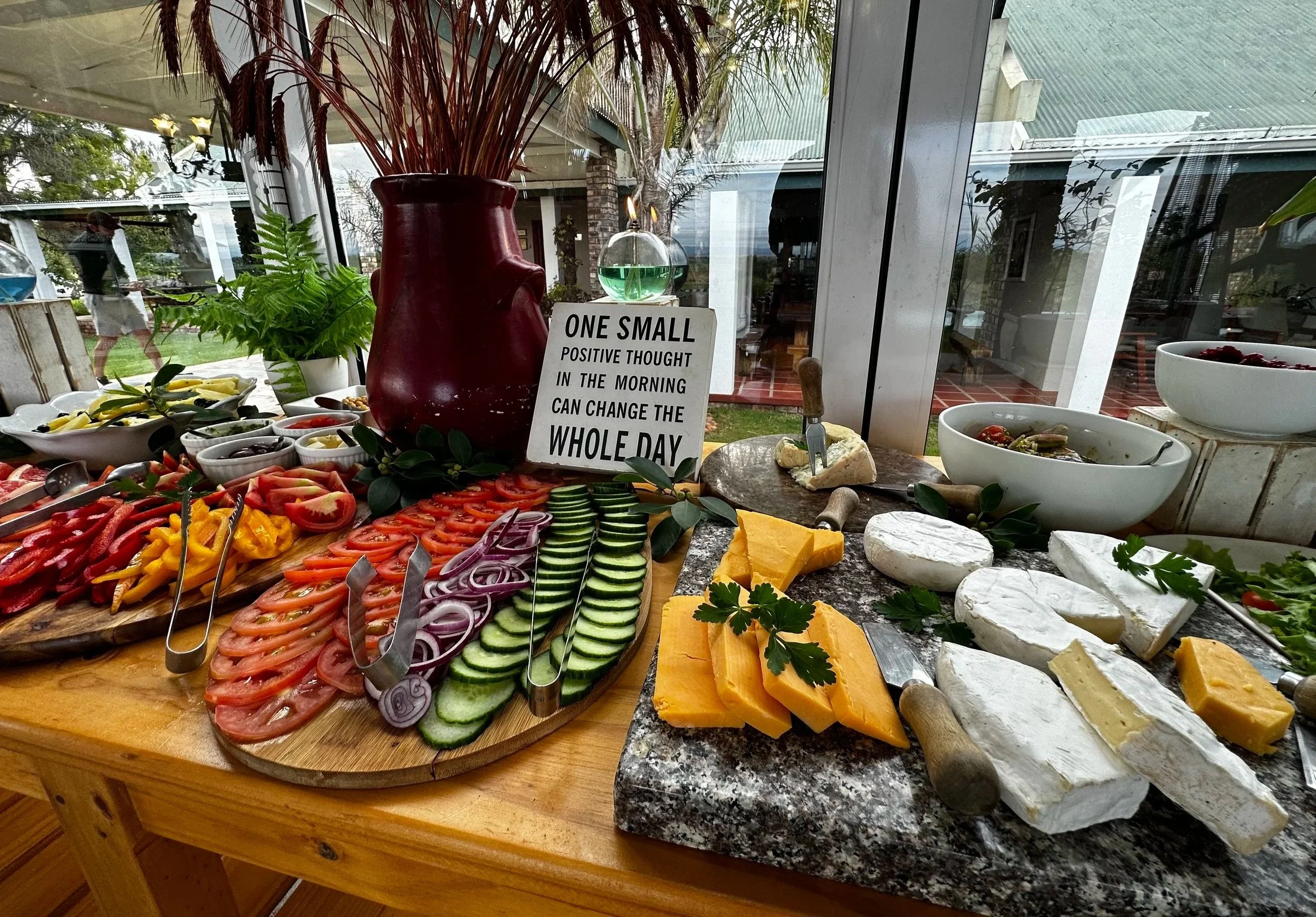 A buffet table with sliced vegetables, cheeses, and dips near a window, with a sign that says, 'One small positive thought in the morning can change the whole day.'