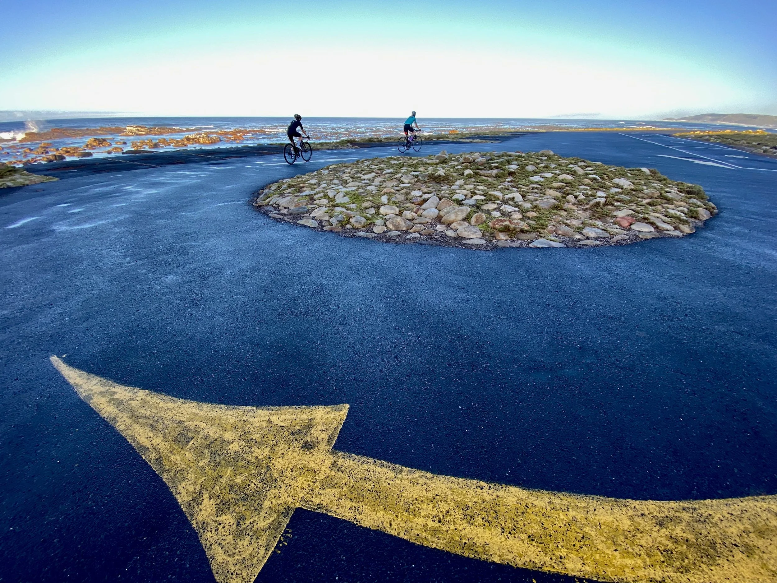 Two cyclists riding on a coastal road with ocean waves and rocky shoreline in the background during daytime.