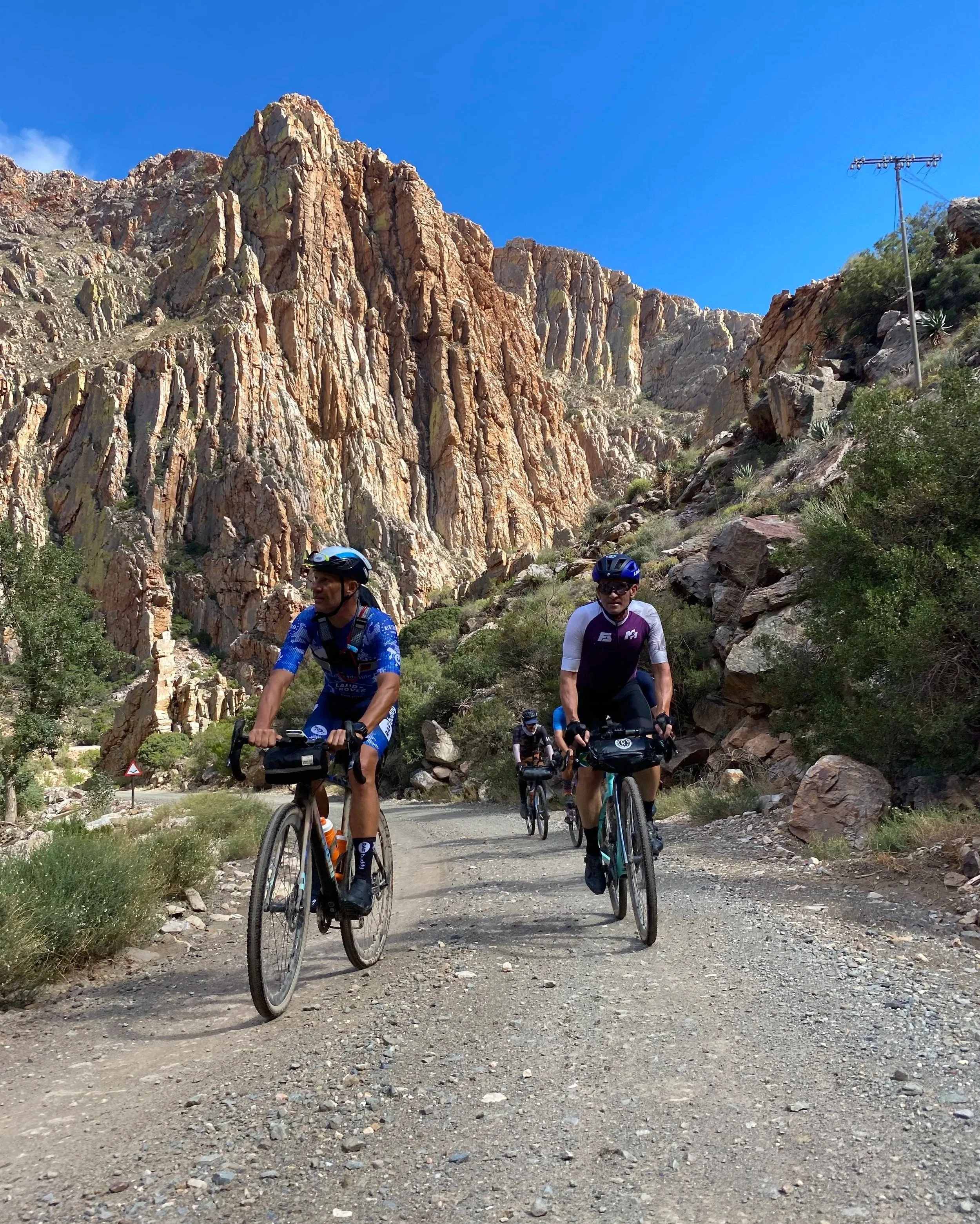 Four cyclists riding on a rocky dirt trail surrounded by desert plants, with a mountain range and clear blue sky in the background.
