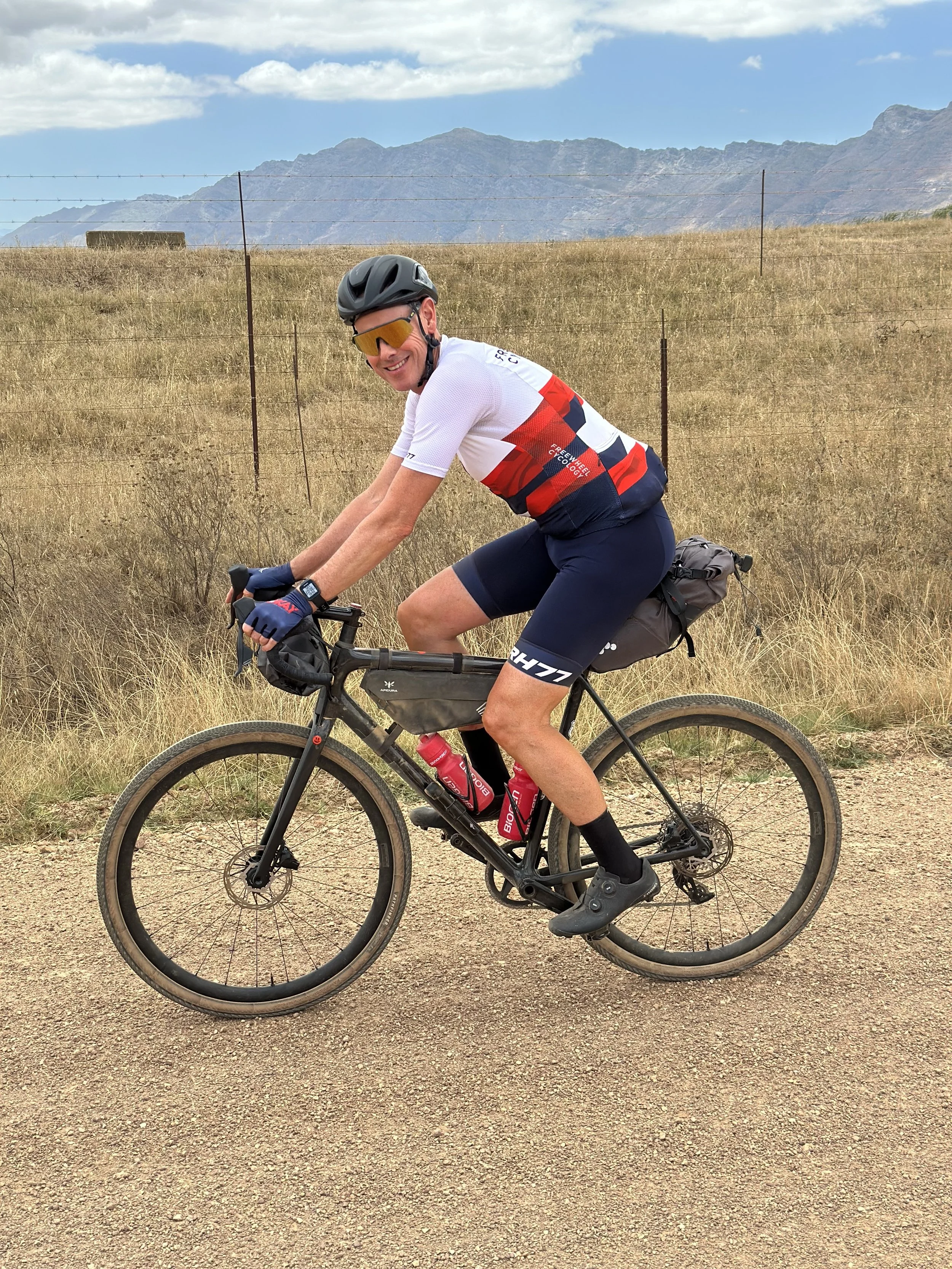 Nic White riding a mountain bike on a dirt trail in a dry, grassy field with mountains in the background and a blue sky with clouds.