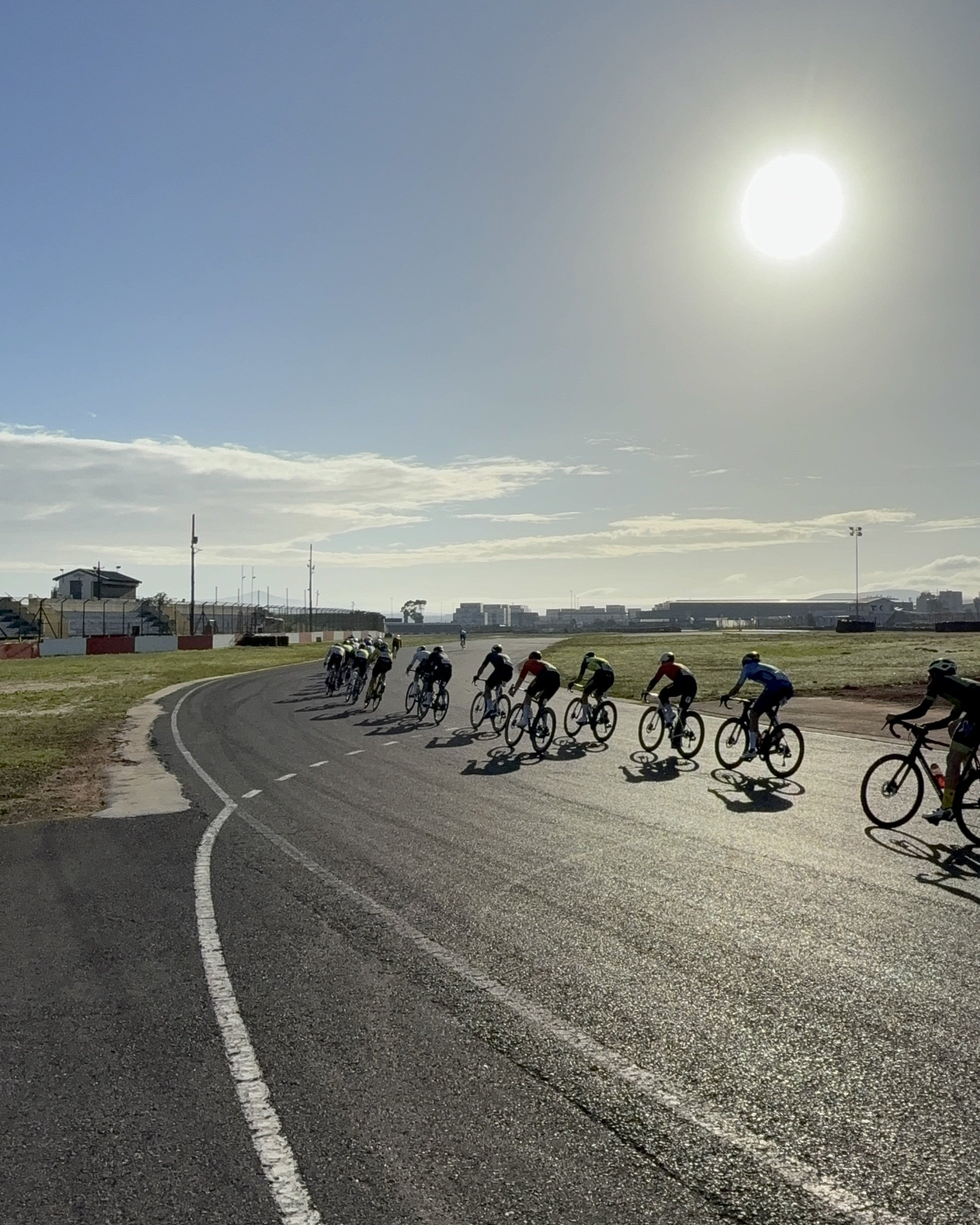 A group of elite cyclists racing on the Killarney race track beneath a bright sky with the sun shining.