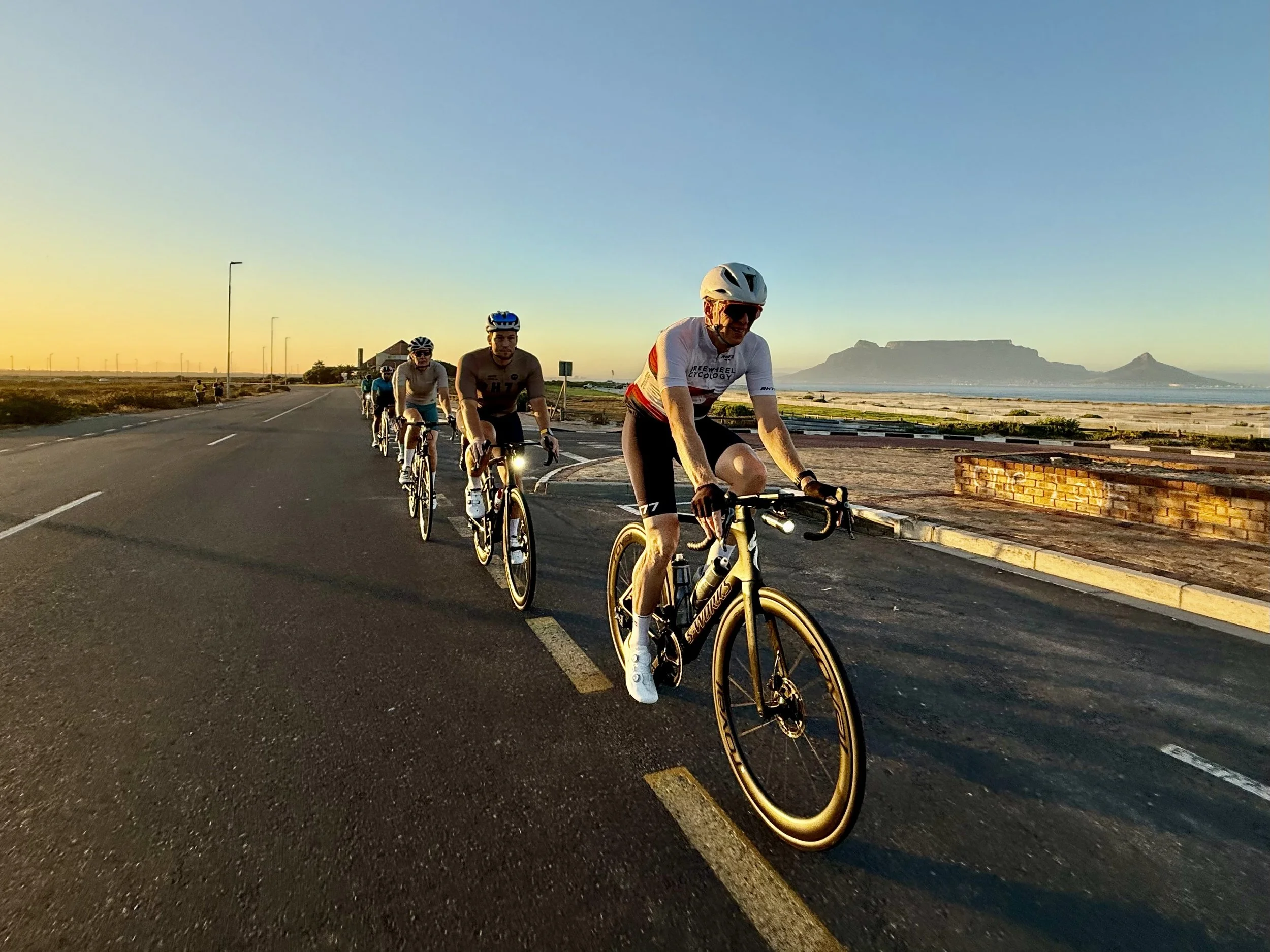 A group of cyclists riding along a road at sunset with mountains in the background.